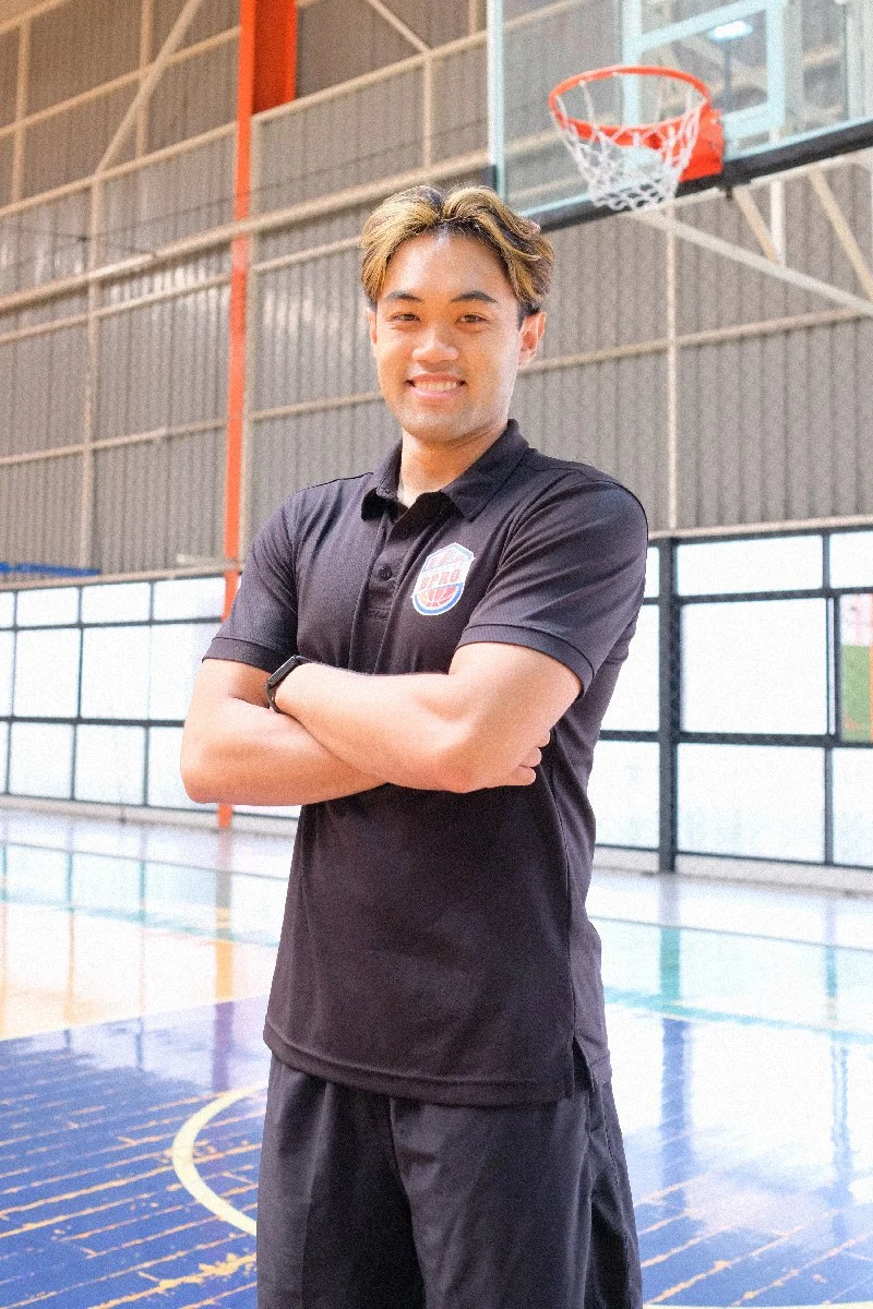 A young man standing with arms crossed in an indoor basketball court, smiling, with a basketball hoop above.