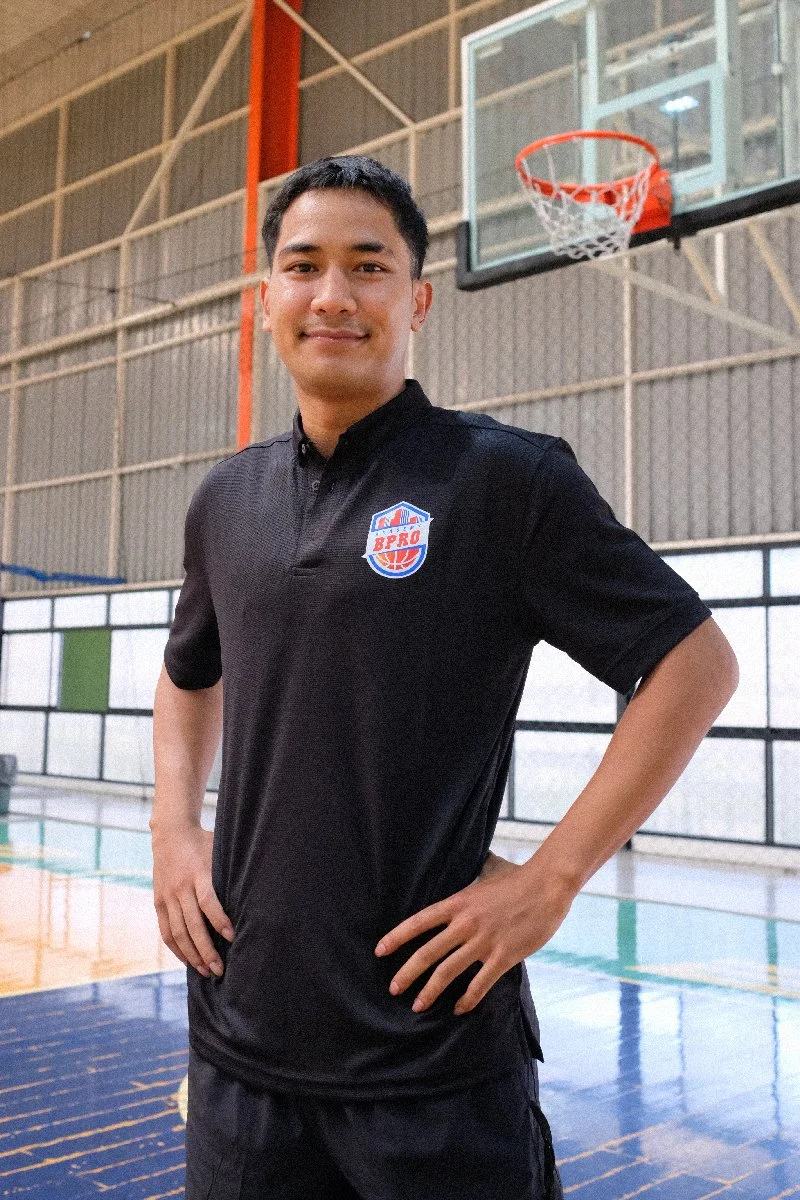 Young man in a black sports shirt standing in an indoor basketball court, smiling, with a basketball hoop in the background.