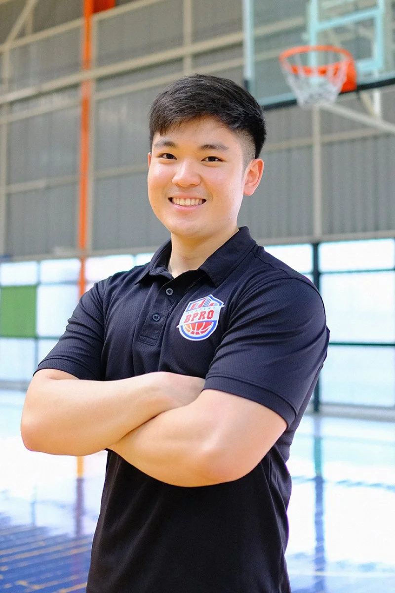 Young man smiling with arms crossed standing in an indoor basketball court, wearing a black polo shirt with a sports logo.