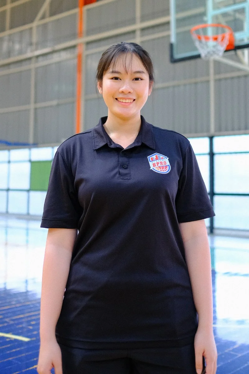 A young woman wearing a black basketball uniform with a logo that reads 'BPRO' stands in an indoor basketball court, smiling at the camera.