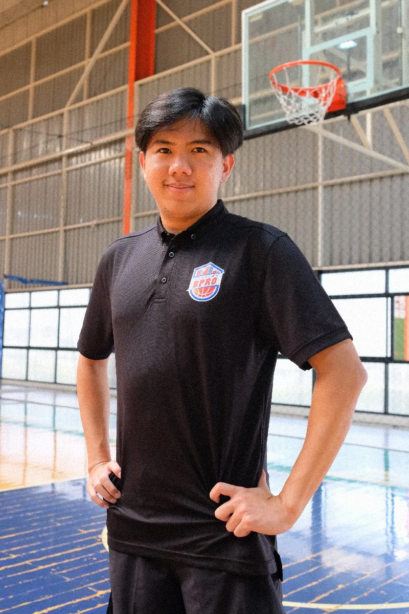 A young man standing confidently in an indoor basketball court, wearing a black polo shirt with a basketball logo on the chest, with one hand on his hip and the basketball hoop visible behind him.