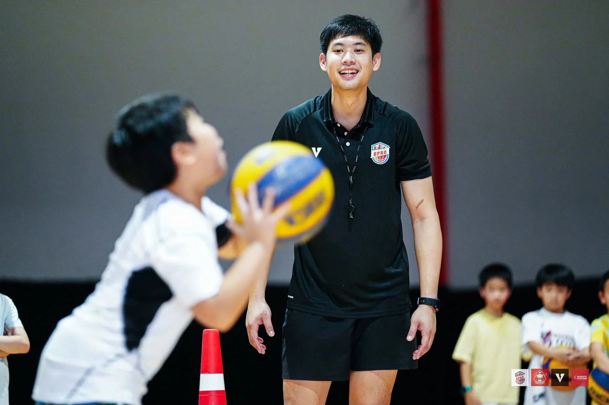 A coach or instructor supervising a young boy during a volleyball drill in a gym. The boy is holding a volleyball and the coach is observing him attentively. Several children are seen in the background waiting their turn.