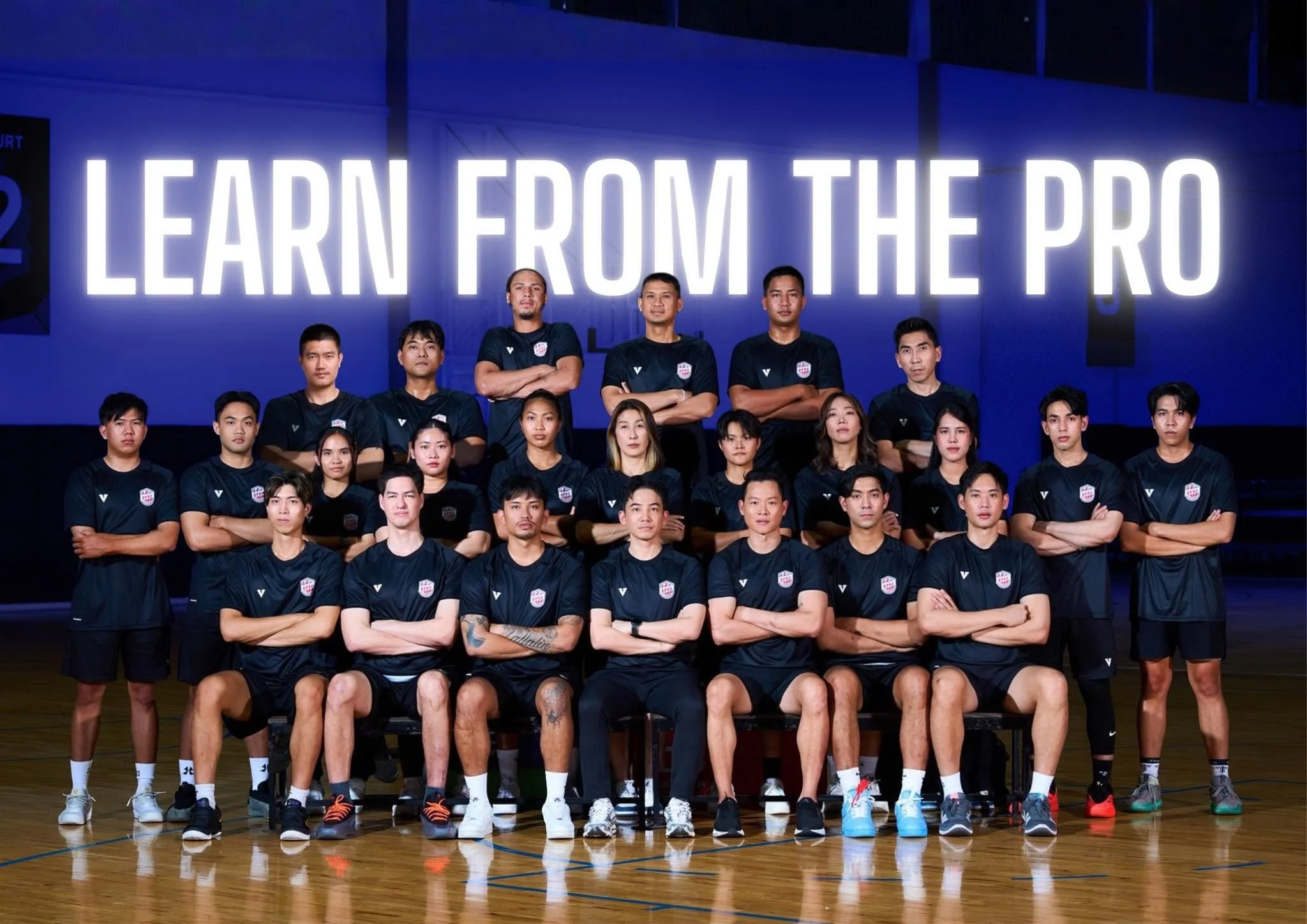 A group of people in sportswear posing for a photo in a gym. They are standing on a basketball court with a large sign behind them that reads 'LEARN FROM THE PRO.'