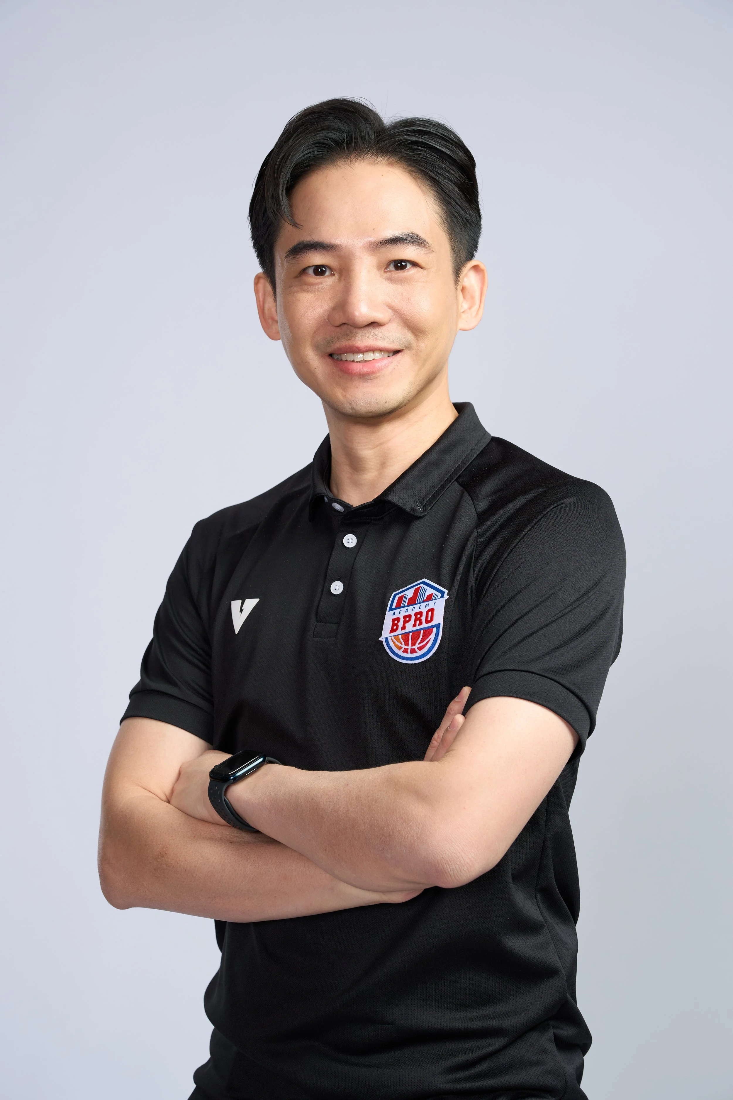 A young man with black hair, wearing a black polo shirt with a sports logo and an Apple Watch, standing against a light gray background, smiling, with his arms crossed.