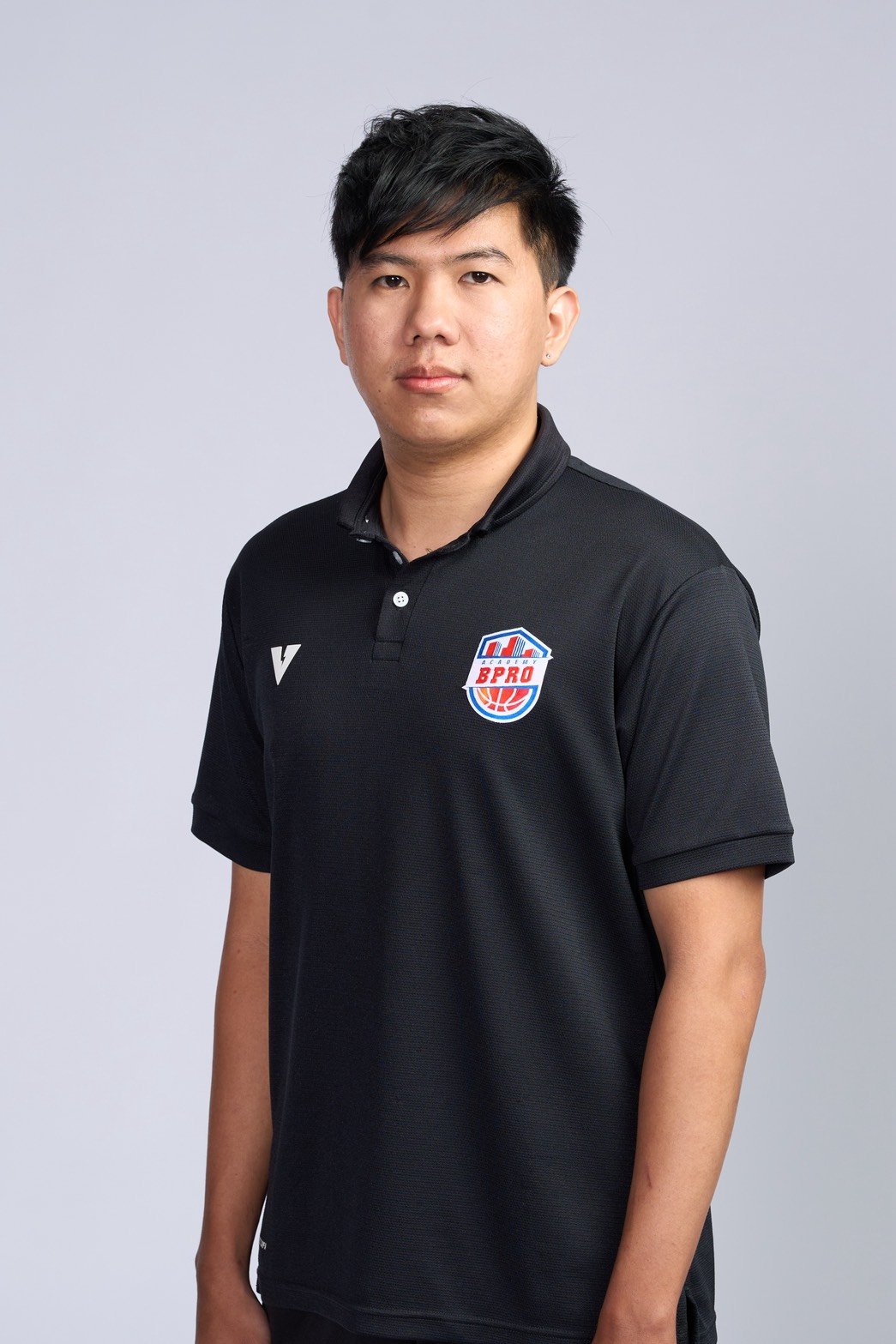 A young man standing confidently in an indoor basketball court, wearing a black polo shirt with a basketball logo on the chest, with one hand on his hip and the basketball hoop visible behind him.