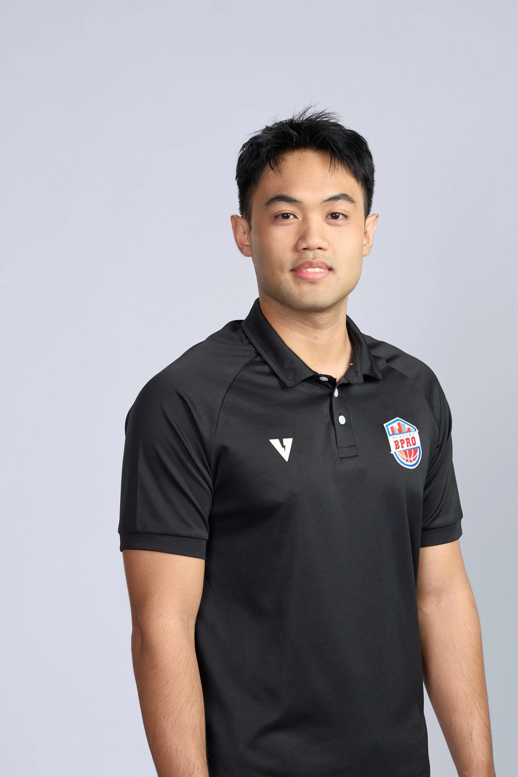 A young man standing with arms crossed in an indoor basketball court, smiling, with a basketball hoop above.