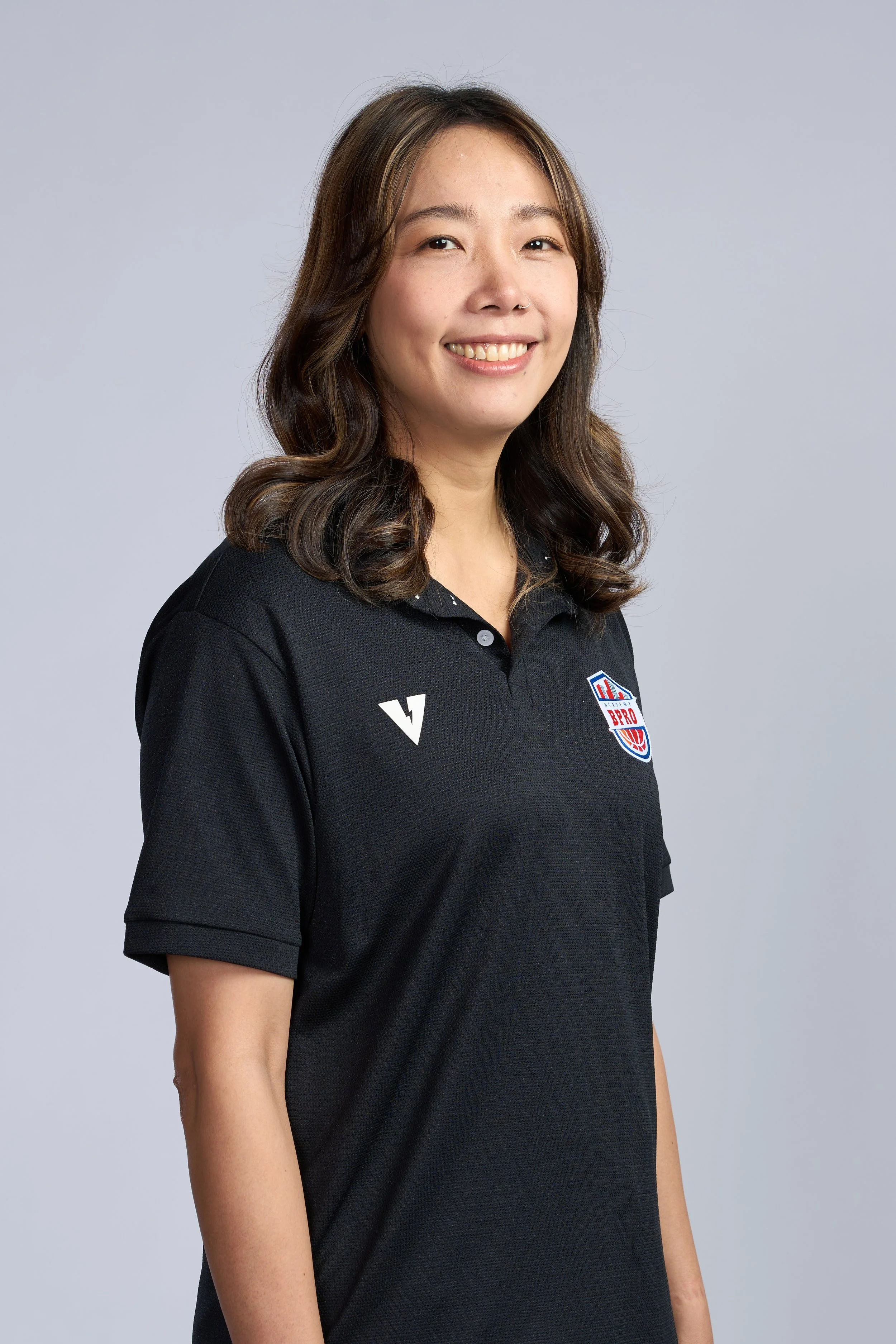 A young woman in a black polo shirt holding a basketball inside a basketball court, smiling at the camera.