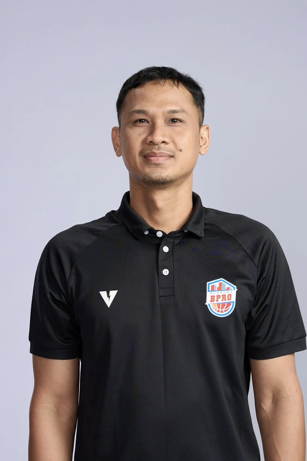 A man in a black polo shirt holding a basketball in a gymnasium with a basketball hoop in the background.