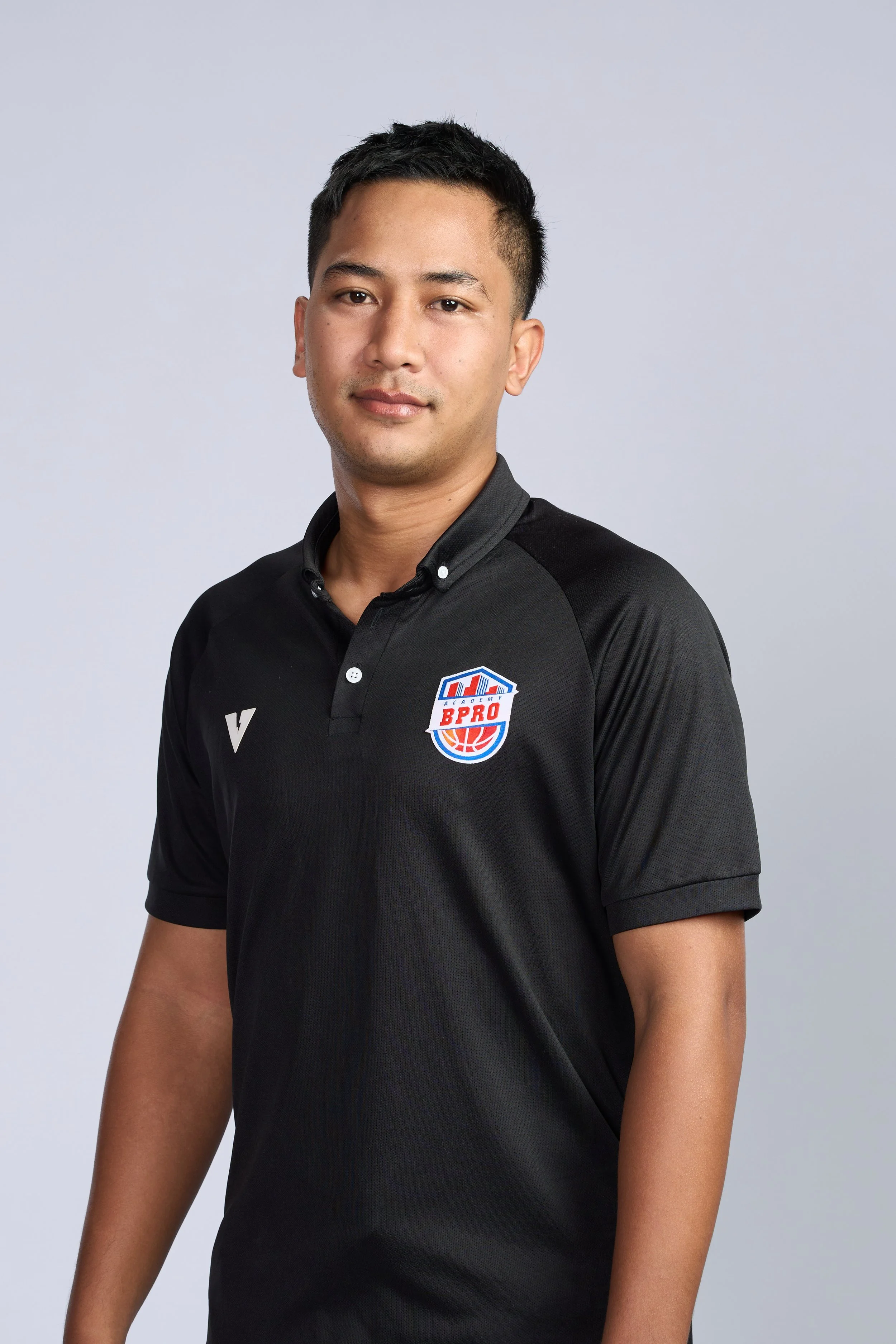 Young man in a black sports shirt standing in an indoor basketball court, smiling, with a basketball hoop in the background.