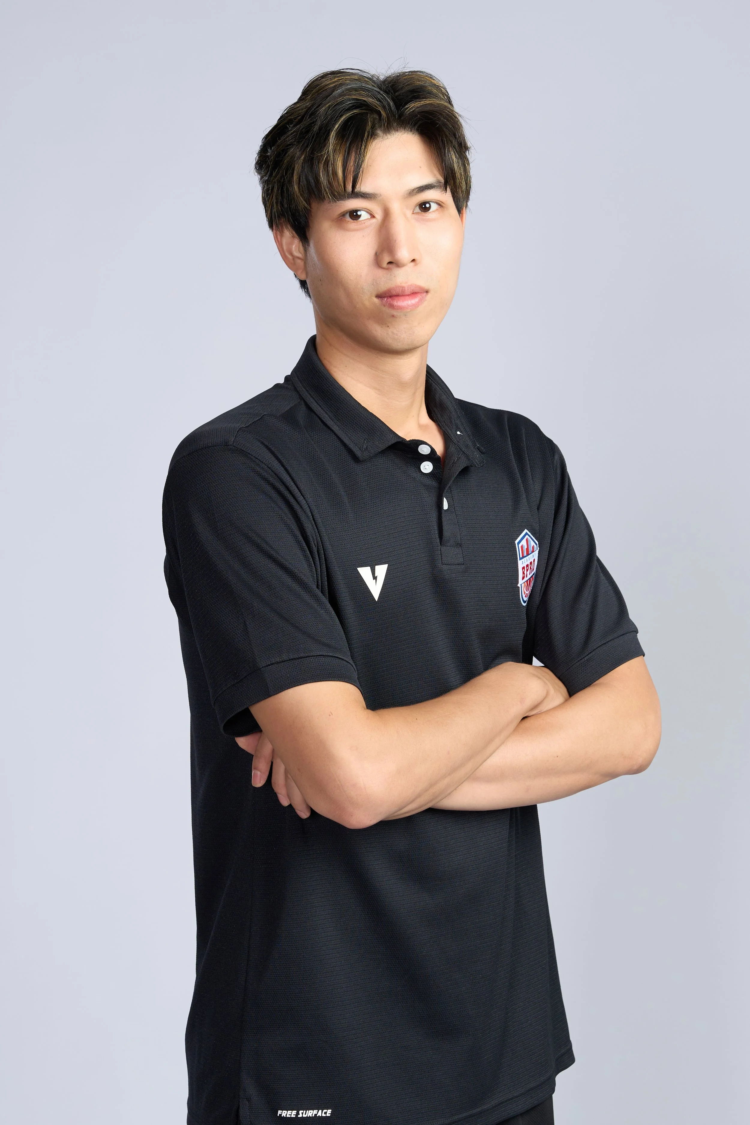 A man standing in a basketball gymnasium holding a basketball with his right hand. He is wearing a black polo shirt with a BPRO logo on it, and his left hand is resting on his hip. The background shows a basketball hoop and a court.