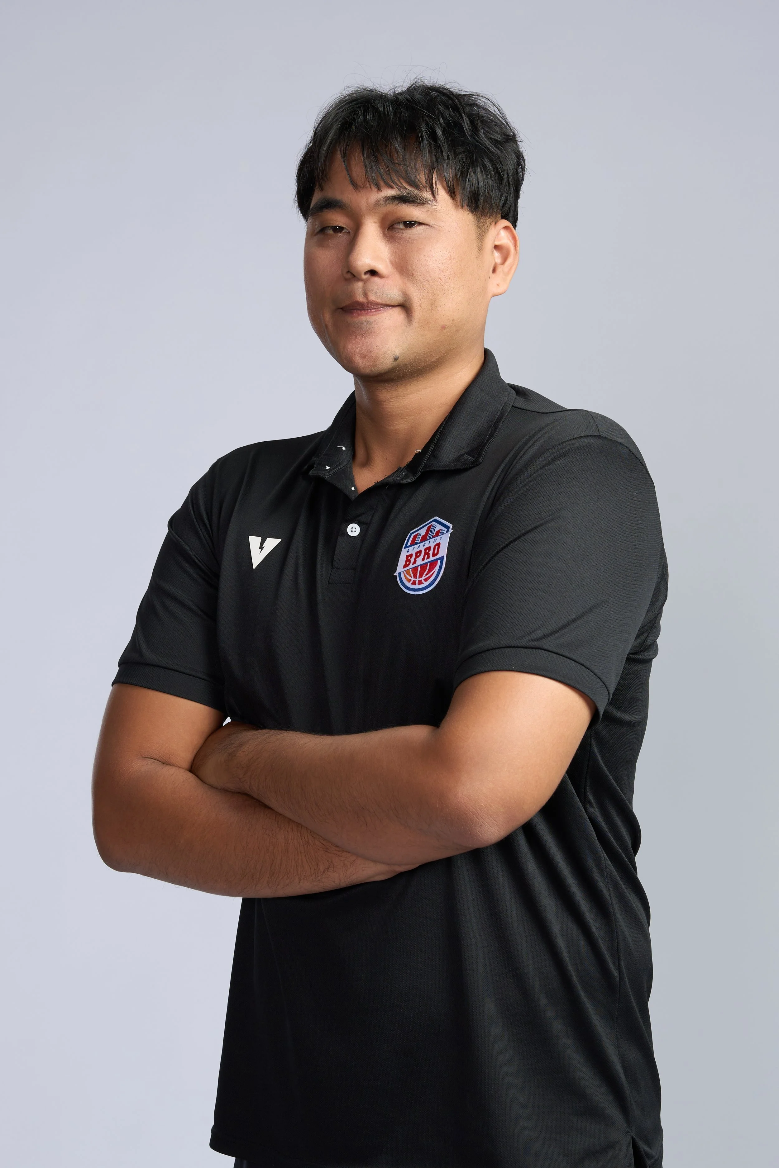A smiling man with crossed arms standing inside a basketball gymnasium. He is wearing a black sports polo shirt with a logo that says 'BPRO' on it. In the background, there is a basketball hoop and part of a basketball court.