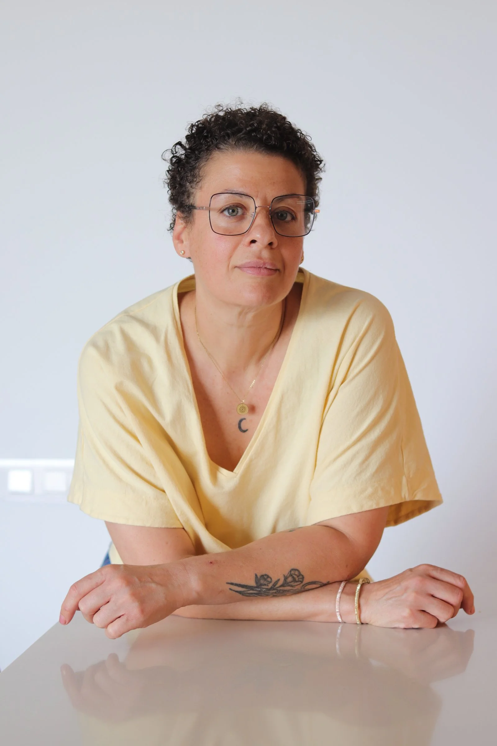 Mujer con cabello rizado, gafas grandes, camiseta amarilla, tatuajes en brazo y collar dorado, sentada en mesa blanca en un fondo blanco.