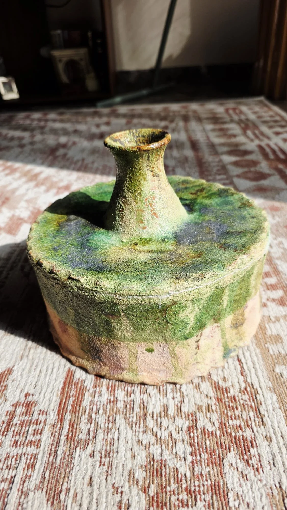 Vaso de cerámica decorativo con superficie áspera, colores verdes y marrón, sobre una alfombra de colores rojo y beige.