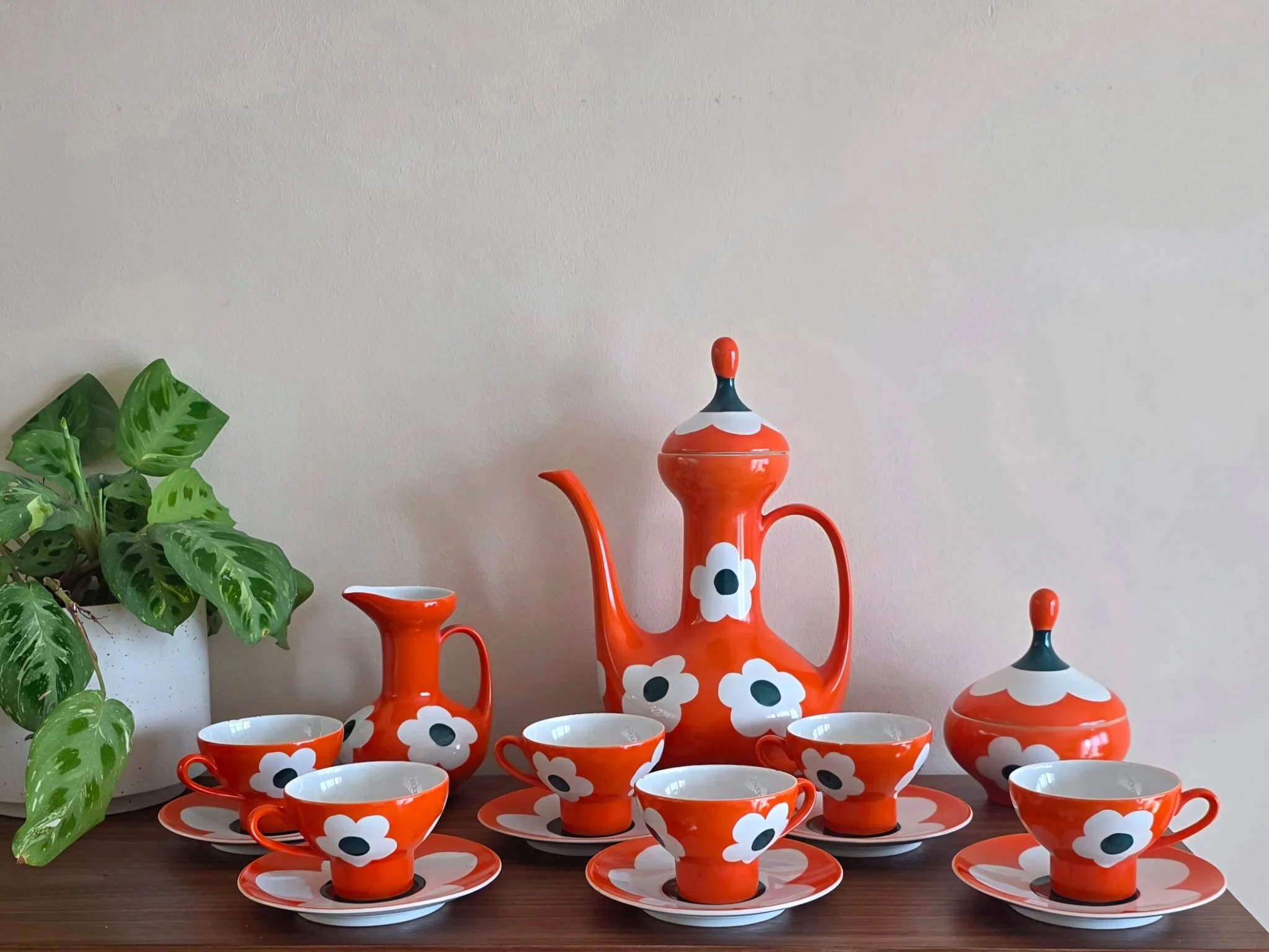 Juego de vajilla decorativa con tazas, platos, una tetera y una tetera más pequeña en color naranja con diseño de flores blancas y puntos oscuros, sobre una mesa de madera, acompañada por una planta de hojas verdes.