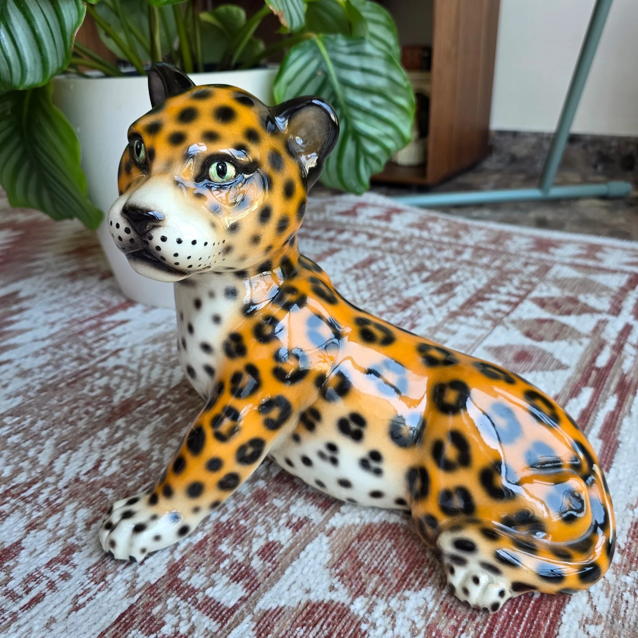 Figura decorativa de un jaguar con ojos grandes, ubicada sobre una alfombra y rodeada de plantas verdes en un ambiente interior.