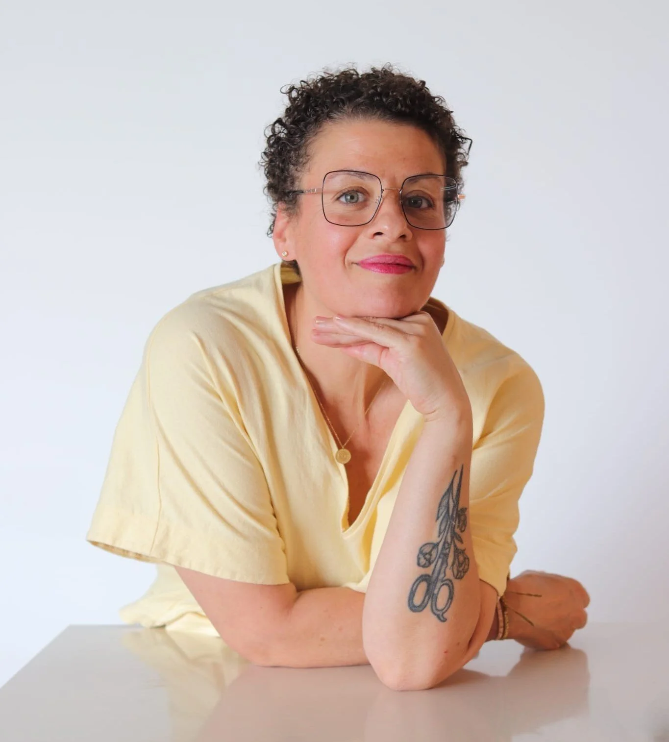 Mujer con cabello rizado, gafas, camiseta amarilla, con tatuajes en el brazo, apoyada con la mano en el rostro, sonriendo suavemente, en un fondo blanco.
