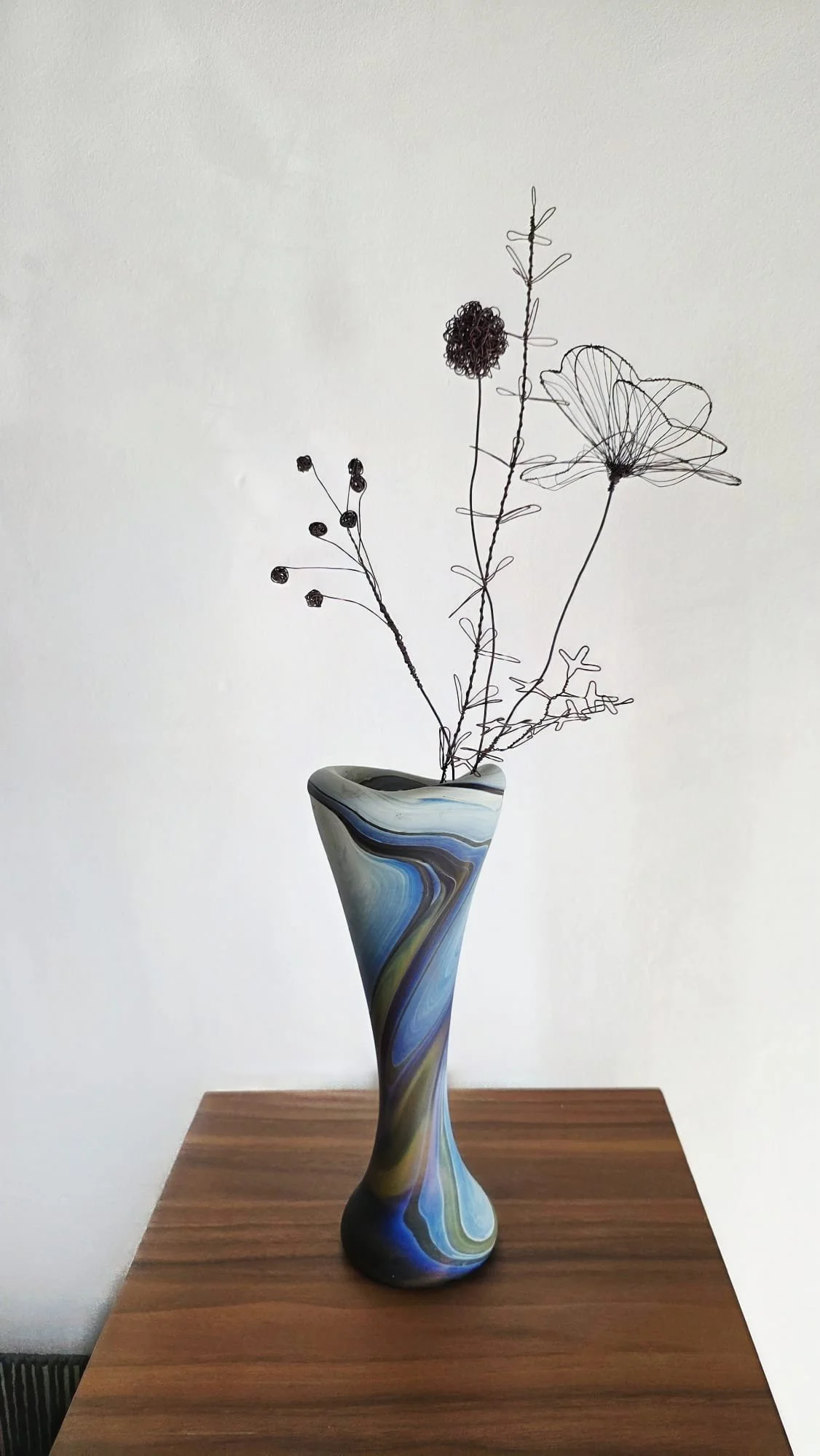 Jarrón de cerámica con diseño multicolor en tonos azules, verdes y grises. Contiene ramas decorativas de metal en forma de flores y hojas, colocadas sobre una mesa de madera.