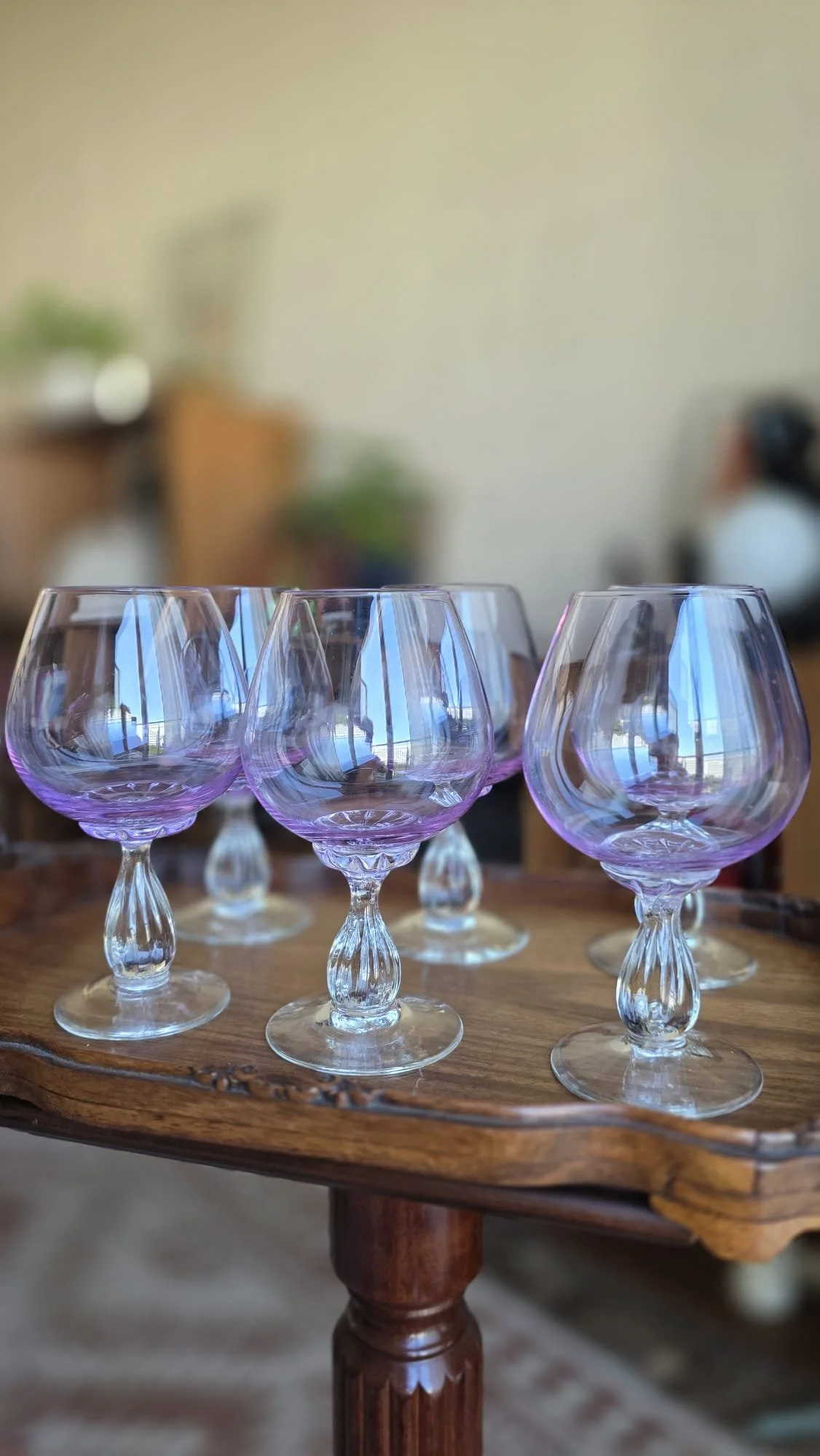 Vasos de cristal con tonalidad violeta en un tablón de madera, en un ambiente interior con muebles y plantas difusos en el fondo.
