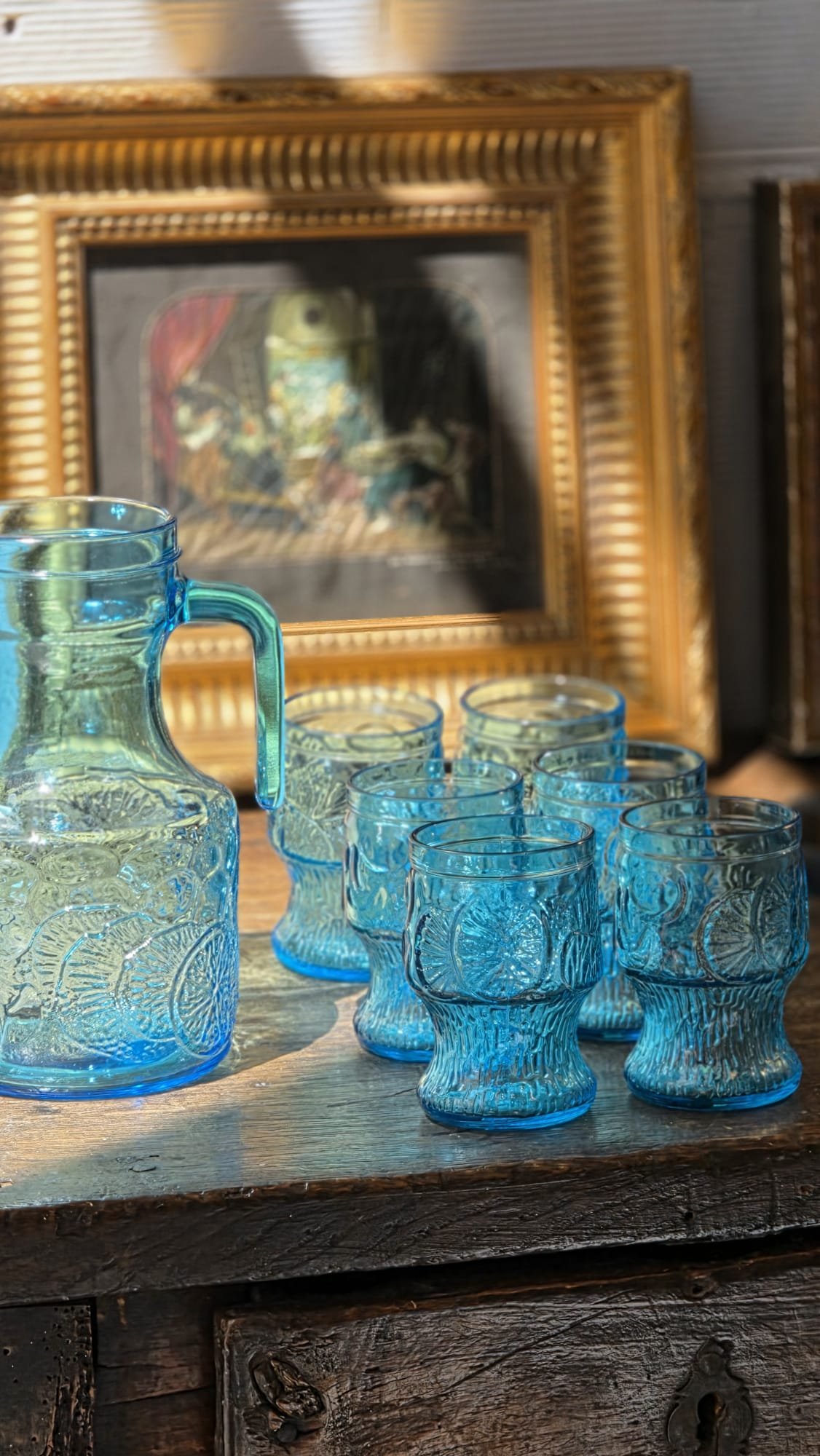 Vasos de vidrio de color azul con grabados, de diferentes tamaños, dispuestos sobre una mesa de madera, con un marco dorado y un cuadro con arte en el fondo.