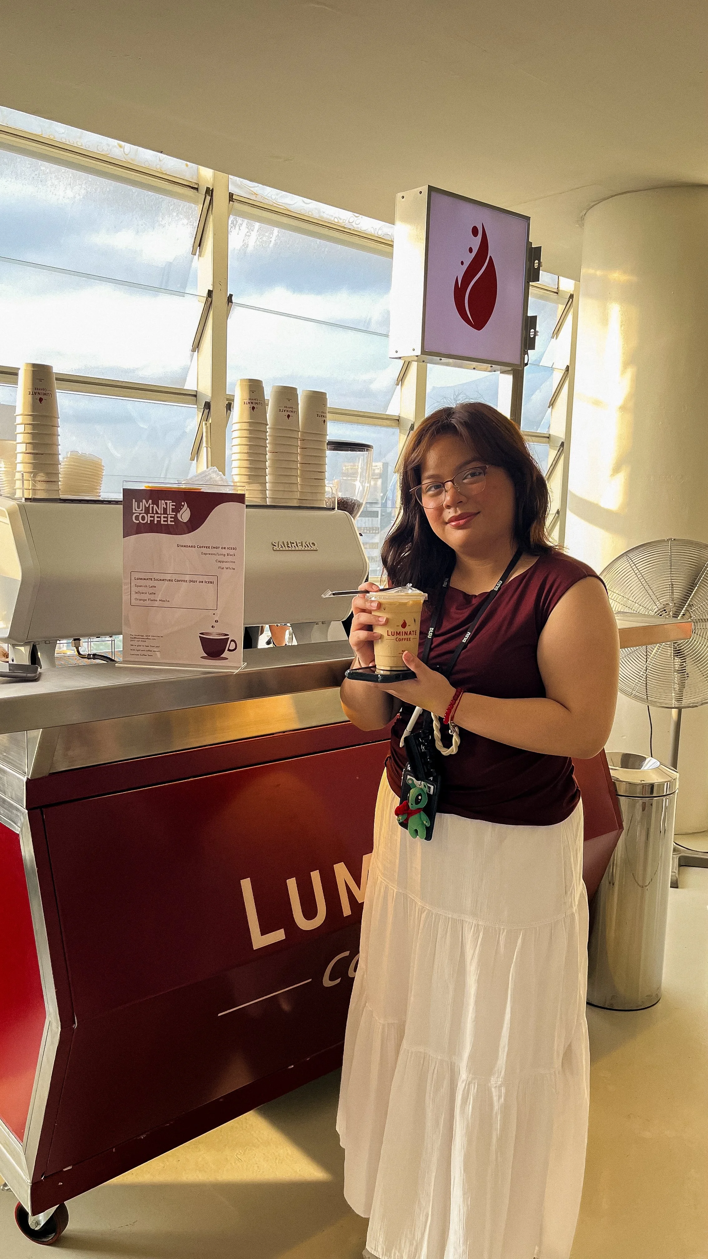 A woman with glasses and wearing a maroon top and white skirt standing at a coffee cart holding a cup of coffee, with a mobile phone and a keychain clipped to her skirt, inside a modern building with large windows.