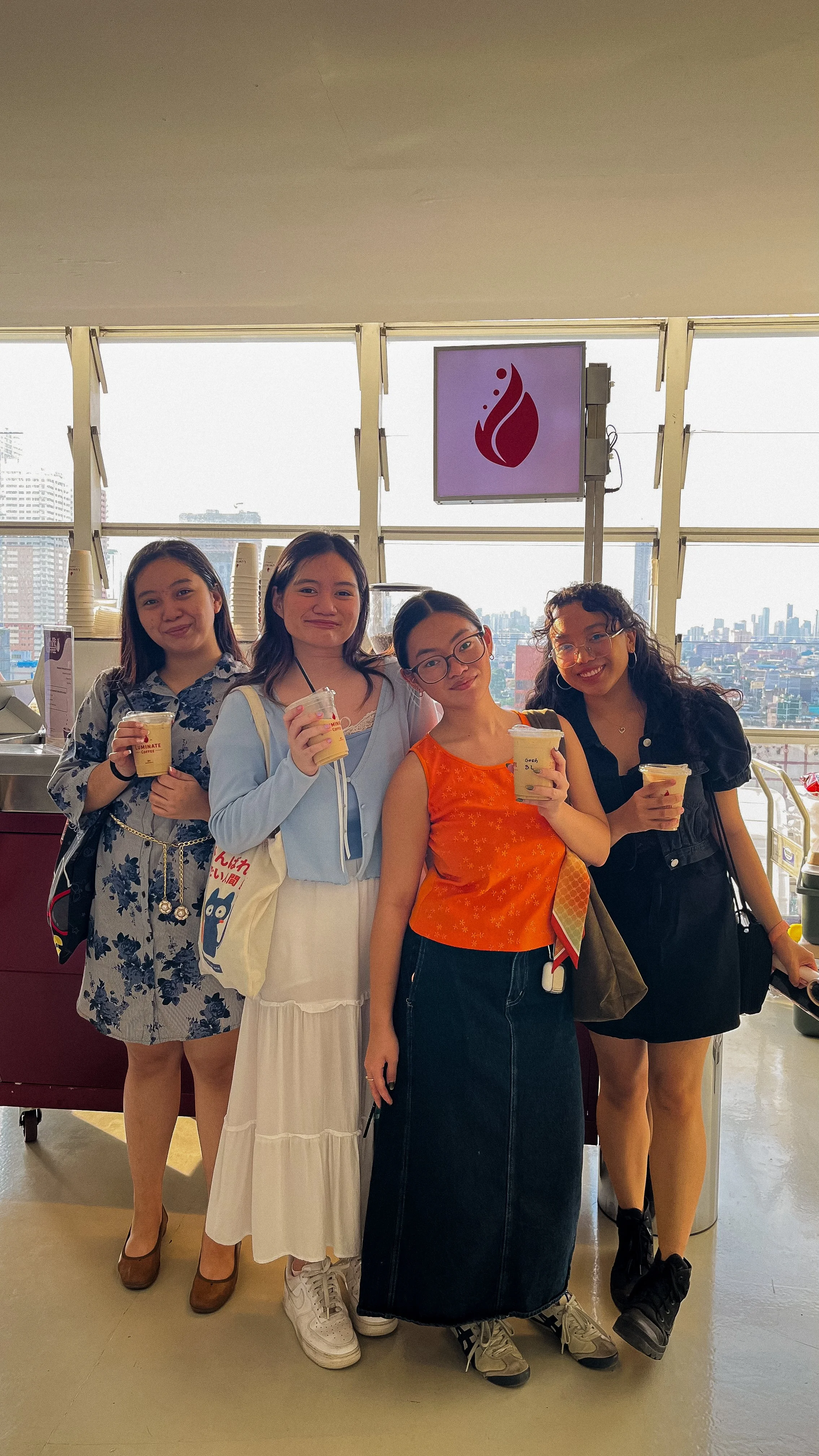 Four young women standing together indoors, holding drinks, with a city skyline visible through large windows behind them.
