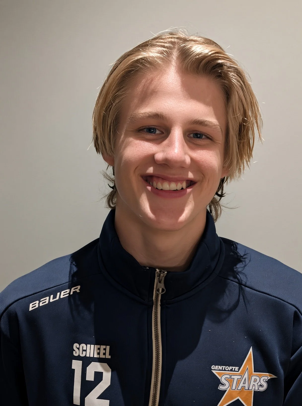 En ung mand med lyst hår, smilende, iført en mørkeblå hockeyjakke med logoet 'Gentofte Stars' og nummer 12 på. Baggrunden er en neutral væg.