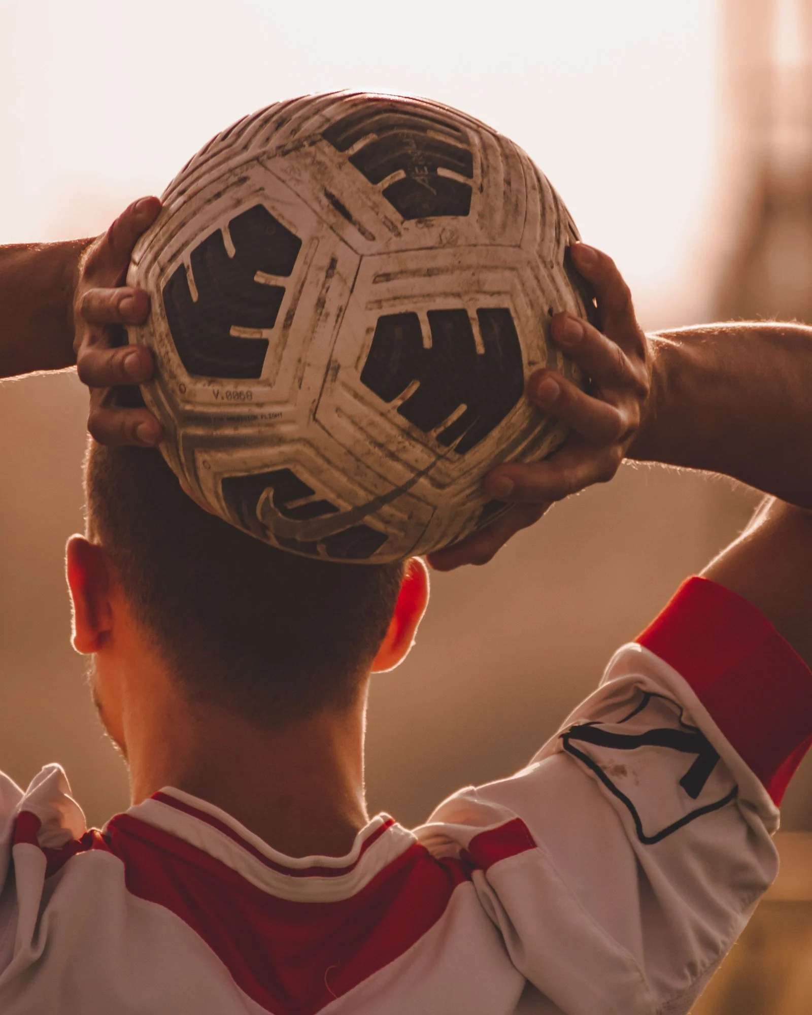 En fodboldspiller set bagfra holder en slidt bold bag hovedet, klar til et indkast, i varmt modlys.