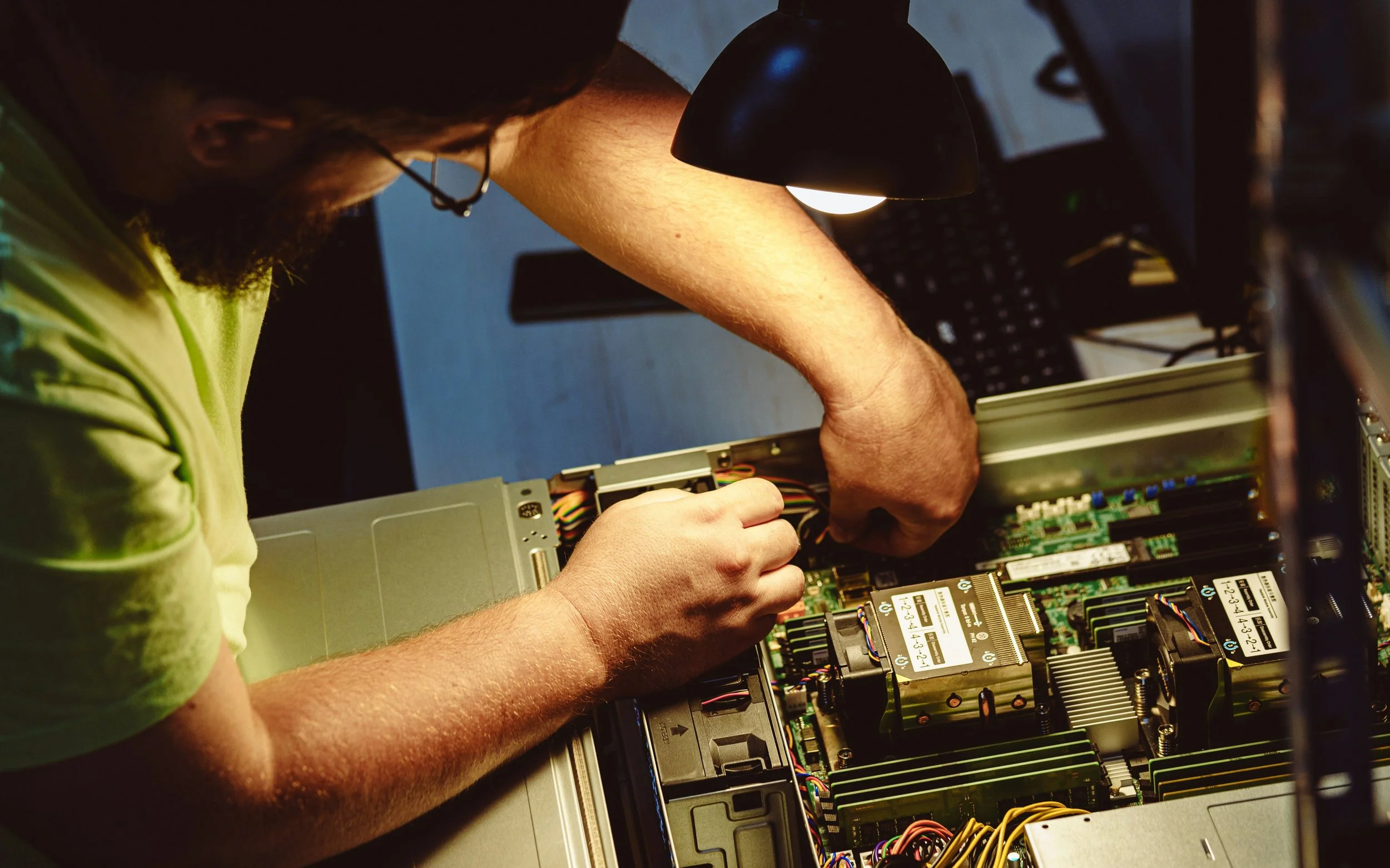 A person working inside a computer case, installing or repairing components with tools. The individual is wearing a neon green shirt and glasses, focused on the hardware.