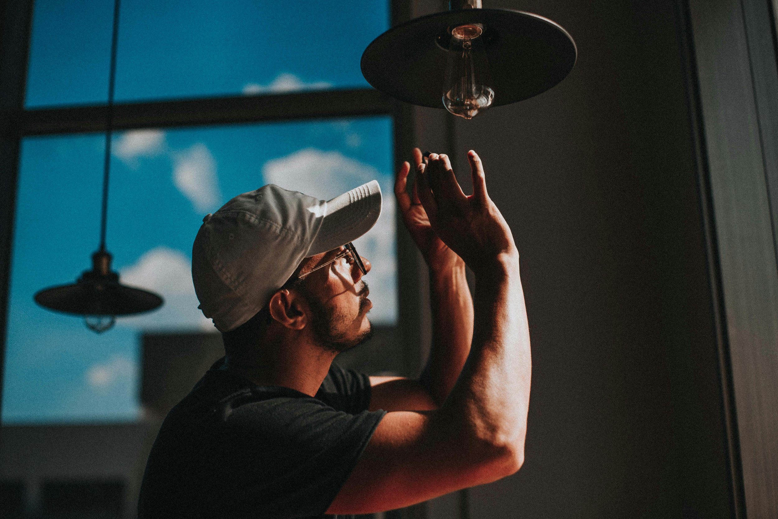 A man wearing a baseball cap and glasses touching a black lampshade; indoor setting with two black pendant lights and a large window showing a cloudy sky.