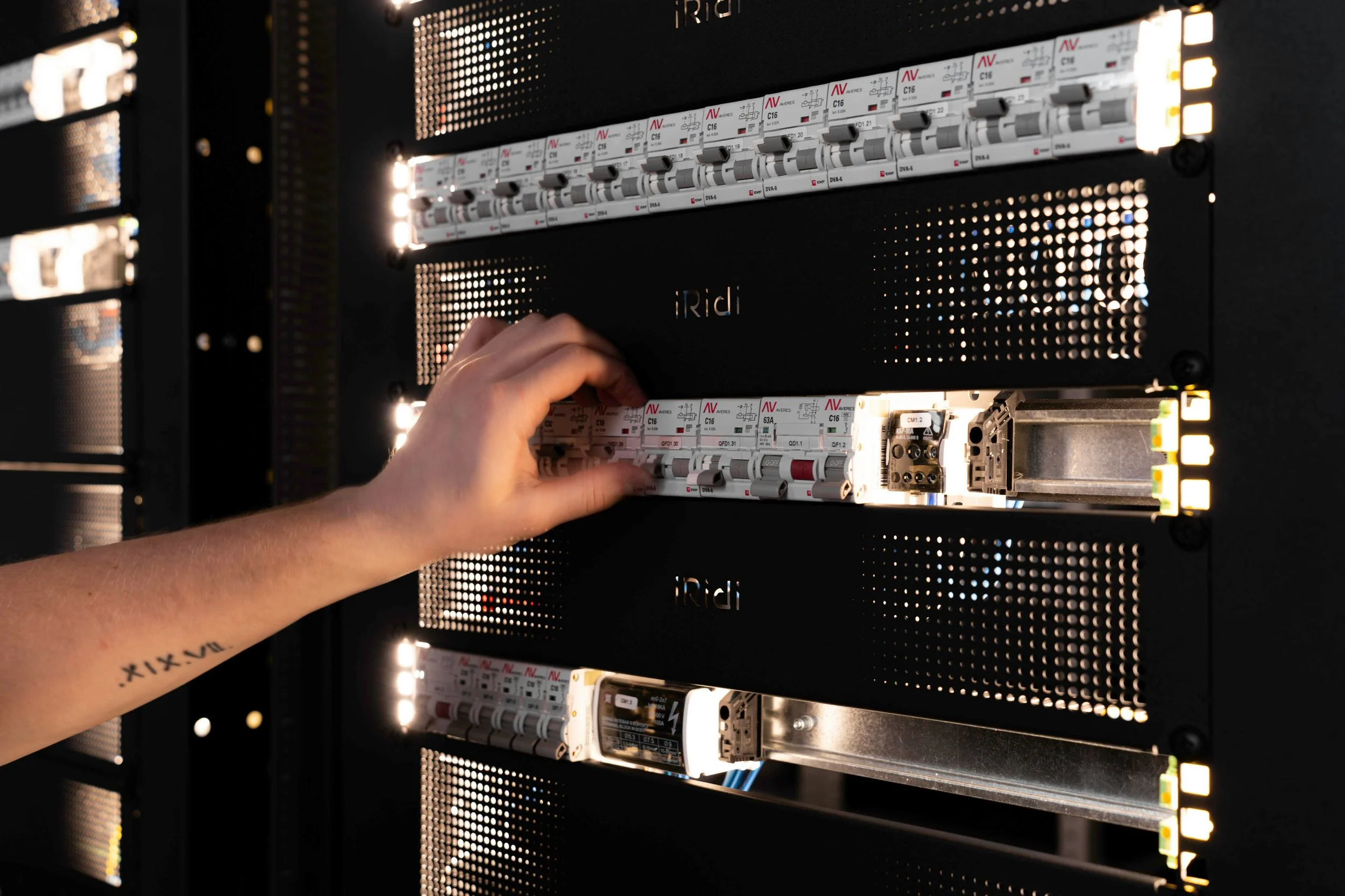 Close-up of an electrical panel with circuit breakers, a person's hand adjusting one of the breakers, and a tattoo on the person's arm.