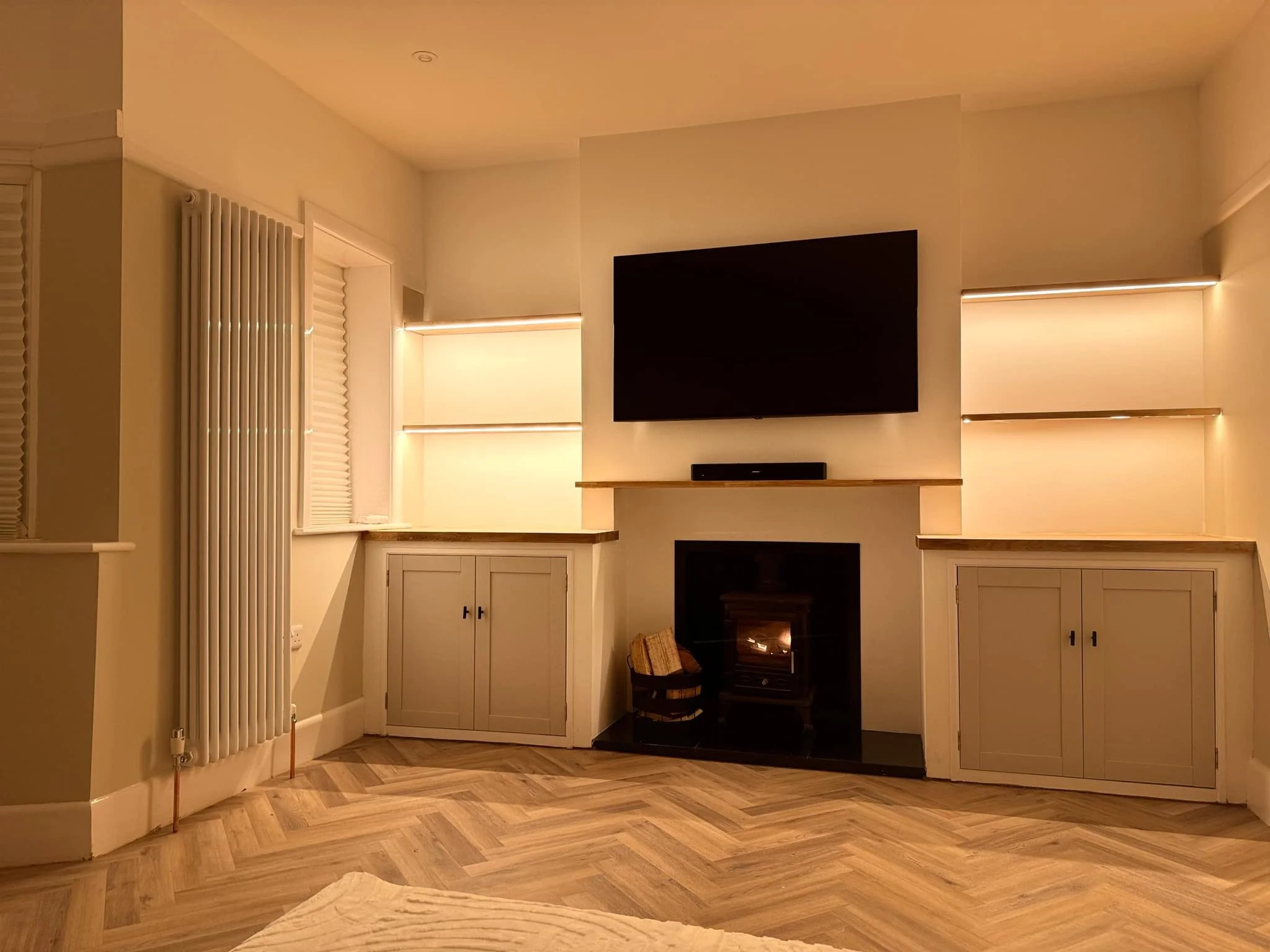 Interior of a living room with a TV mounted on the wall, a fireplace below, beige cabinets, and warm lighting, with wooden flooring.