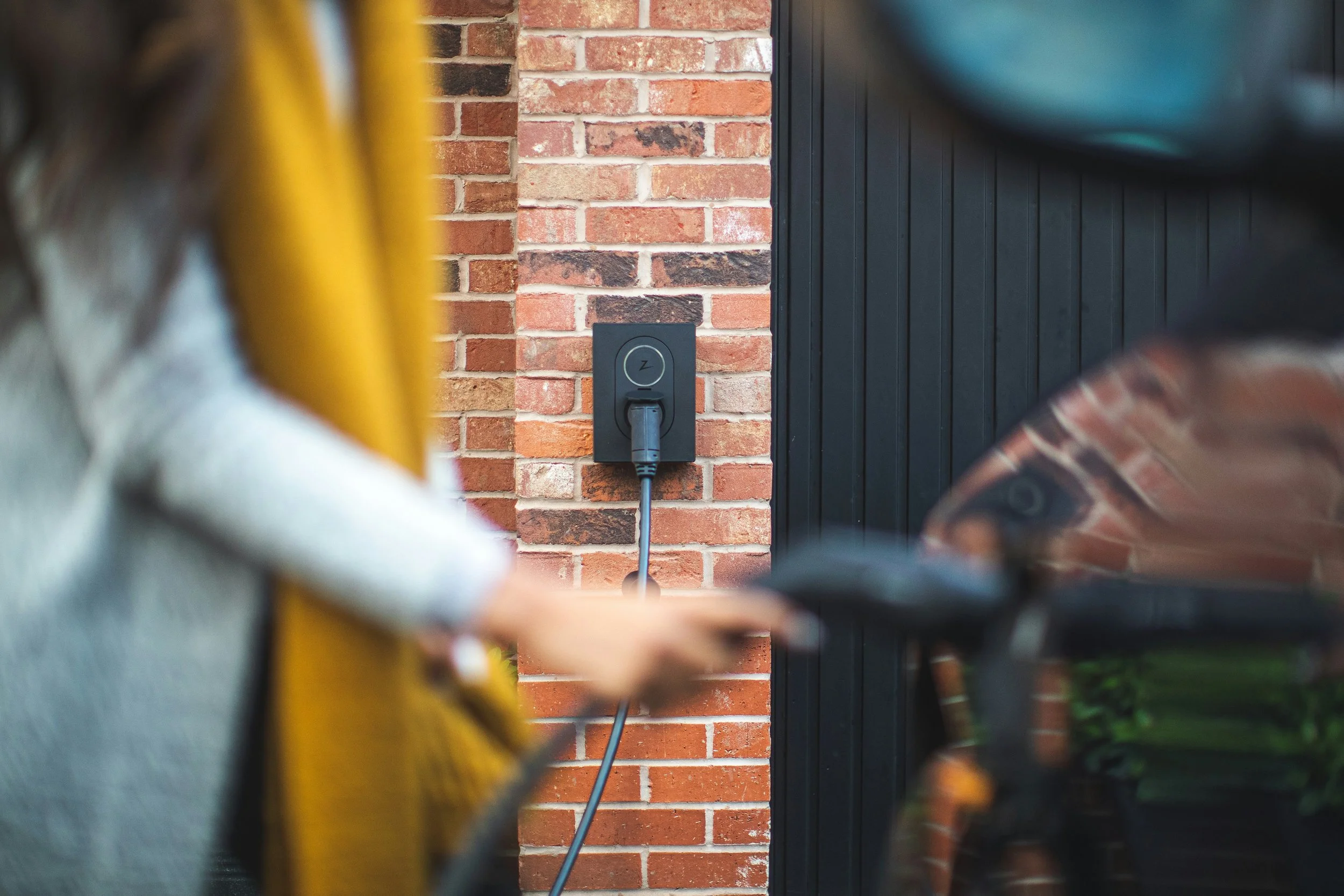 Close-up of two people using electronic devices, with a focus on a brick wall in the background and an electrical outlet on it.