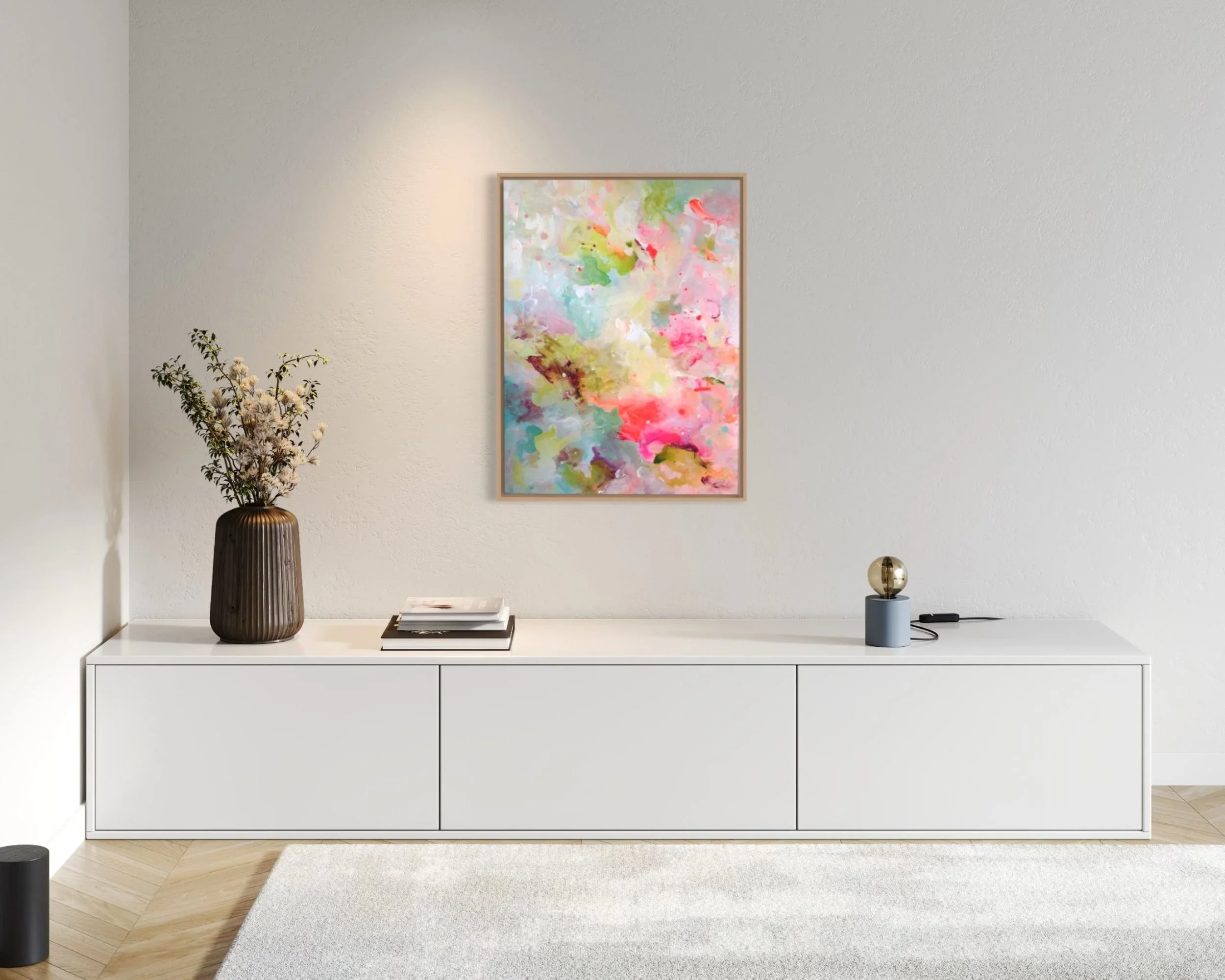 Ein moderner weißer Sideboard mit einer Vase mit getrockneten Blumen, einer kleinen Sammlung von Büchern, einem Bildschirm, einem sphericalle Dekorationsobjekt und einem remote auf der rechten Seite. An der Wand darüber hängt ein buntes abstraktes Gemälde in einem dünnen Holzrahmen.