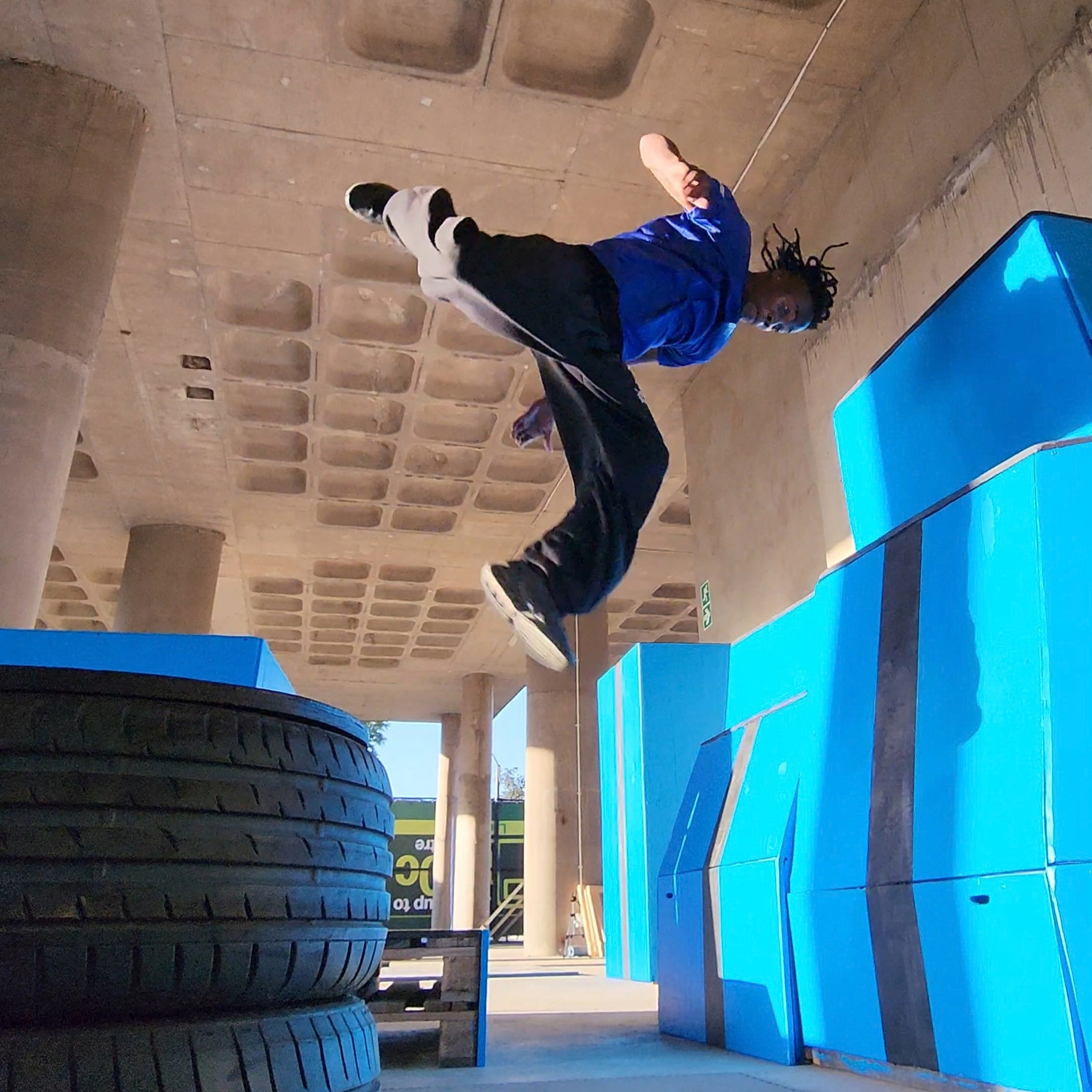 Person in blue shirt and black pants jumping over stacked tires beside a blue wall
