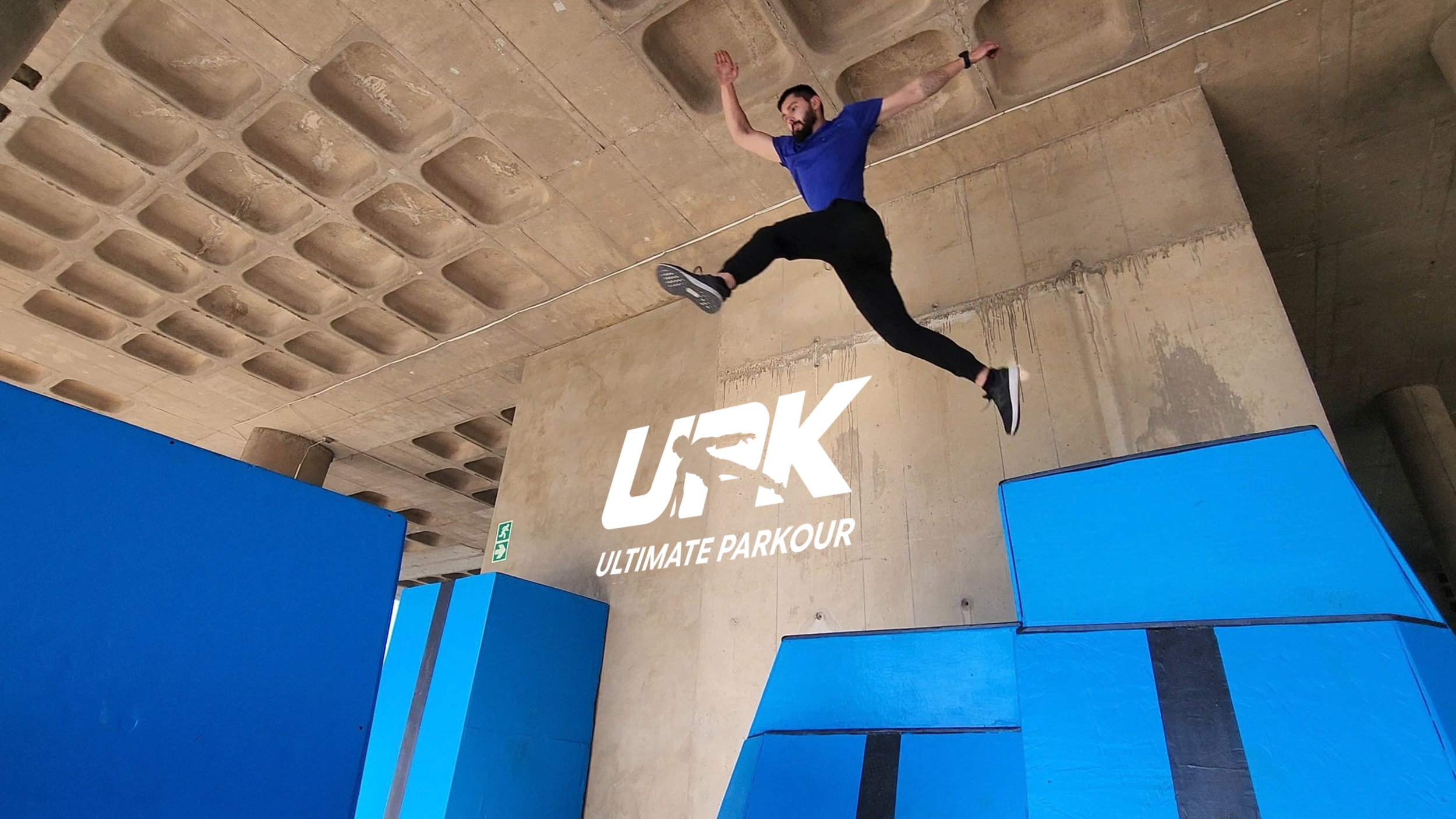 Man performing a parkour jump inside an indoor training facility with blue padded obstacles