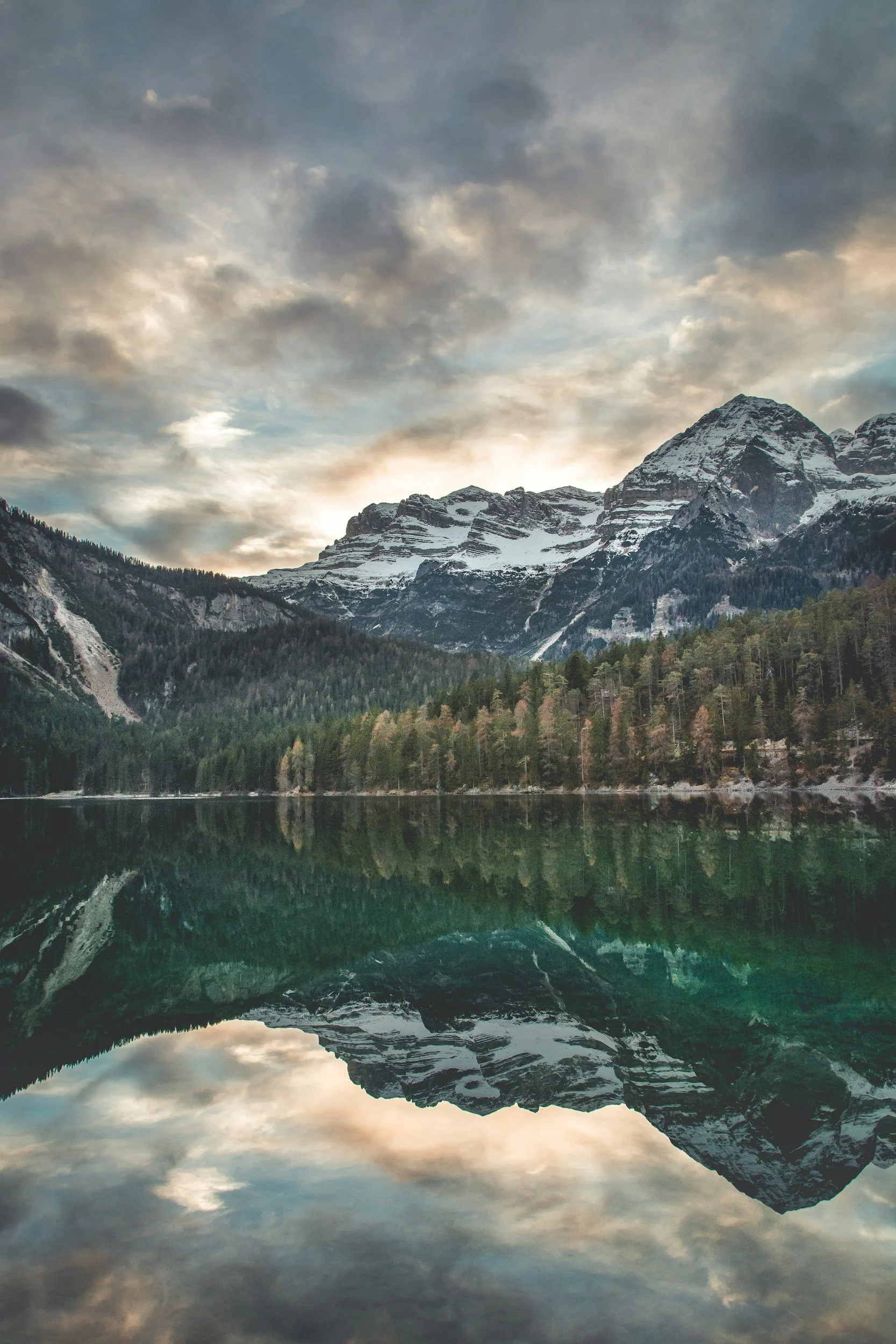 Berglandschaft mit schneebedeckten Gipfeln, einem See im Vordergrund und bewölktem Himmel bei Sonnenuntergang.