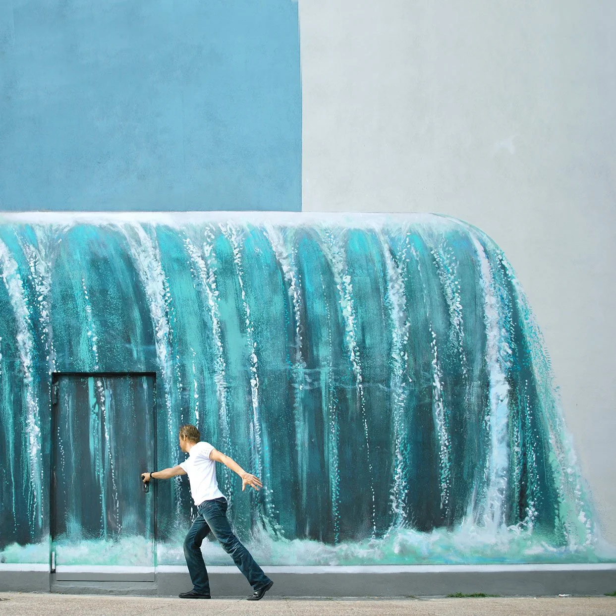 Ein junger Mann in weißem Hemd und dunklen Hosen läuft an einer Wand mit einem Wandbild, das Wasser zeigt, vorbei.