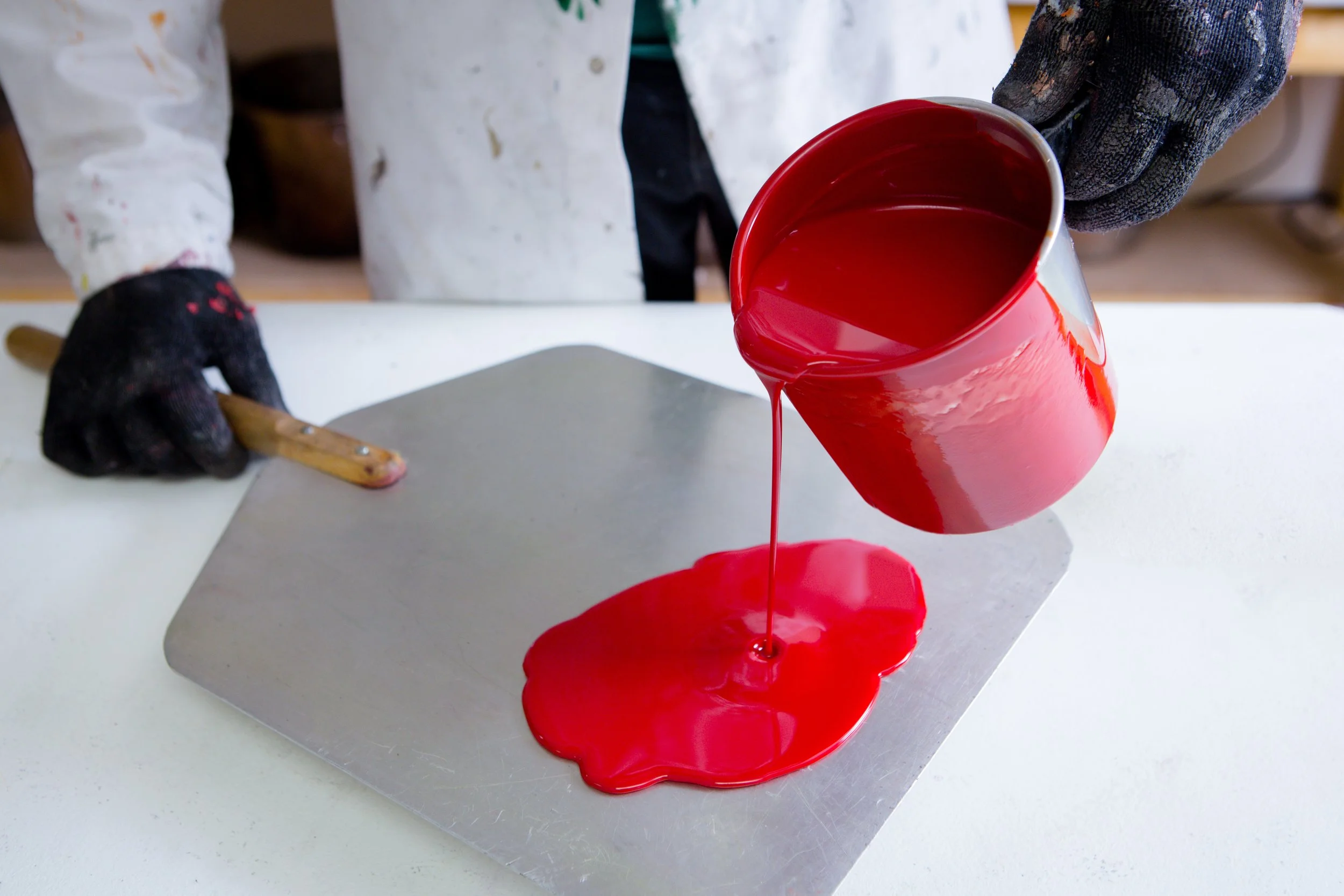 A person wearing black gloves pours red liquid from a red pitcher onto a flat surface.