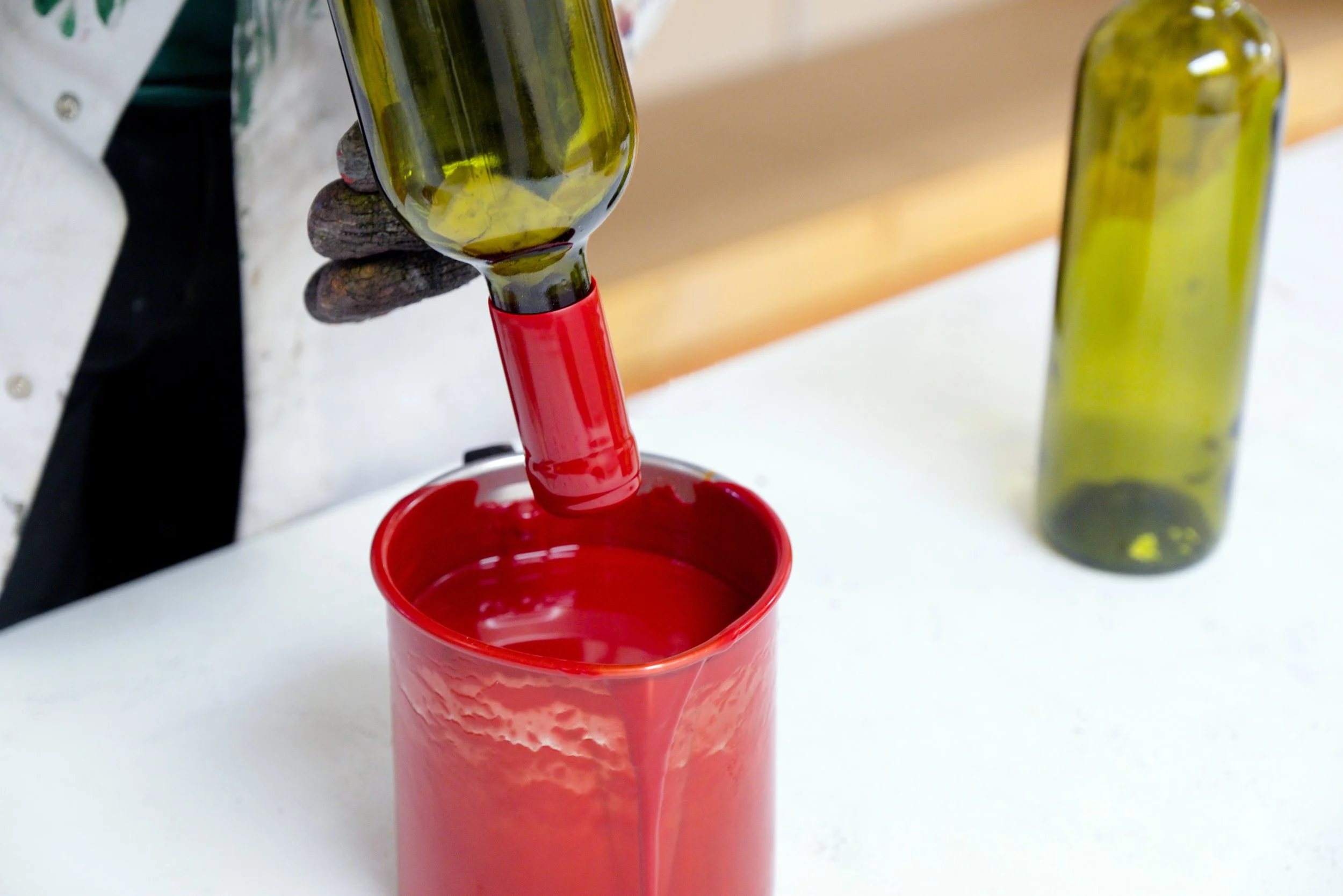 A person's hand holding a green glass bottle over a red plastic cup, with more green glass bottles in the background.