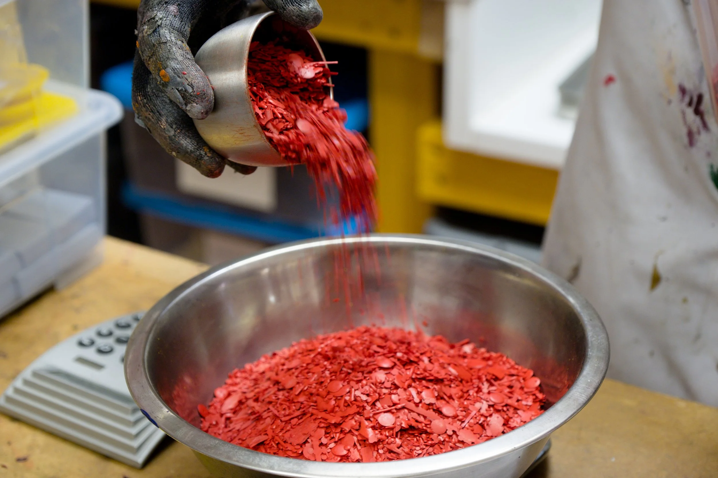 A person with a textured, dirty glove is pouring bright red shattered glass pieces into a metal bowl.