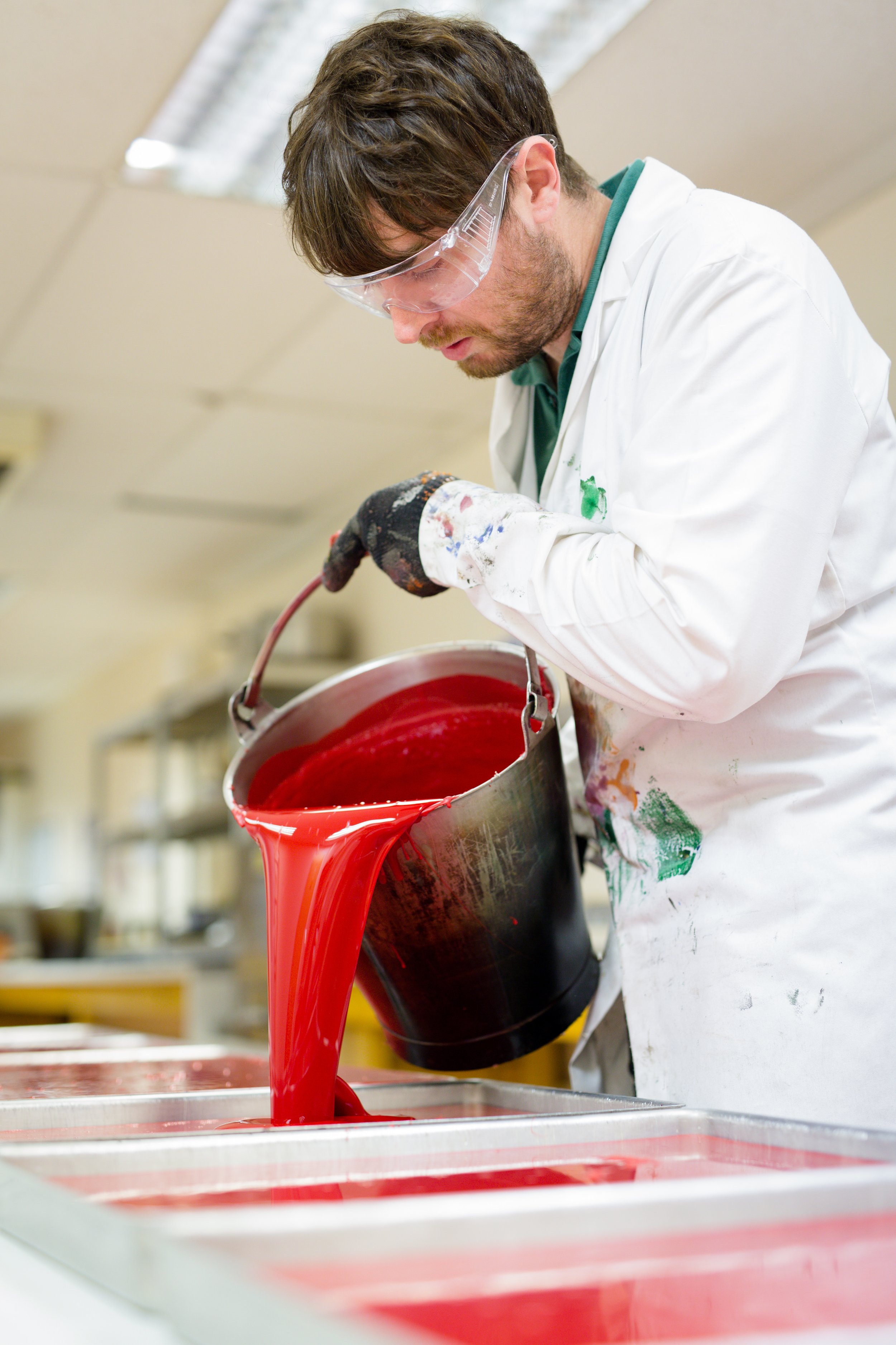 A man wearing safety goggles and a paint-splattered white apron pours red liquid dye into a rectangular container in a workshop.