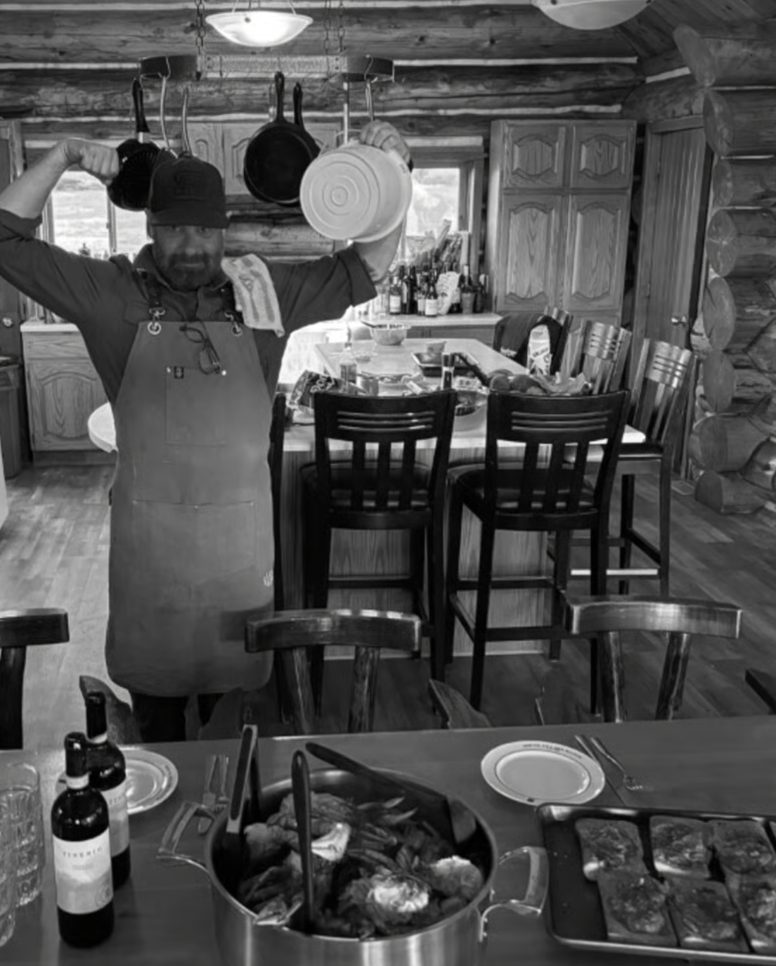 A man in a kitchen holding two pots while flexing his arms. The kitchen has wooden logs, a dining table with chairs, and various kitchen items including bottles and food.