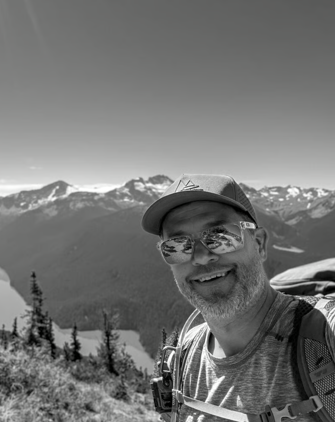 A smiling man with sunglasses and a cap, taking a selfie outdoors on a mountain trail with snow-capped peaks in the background.