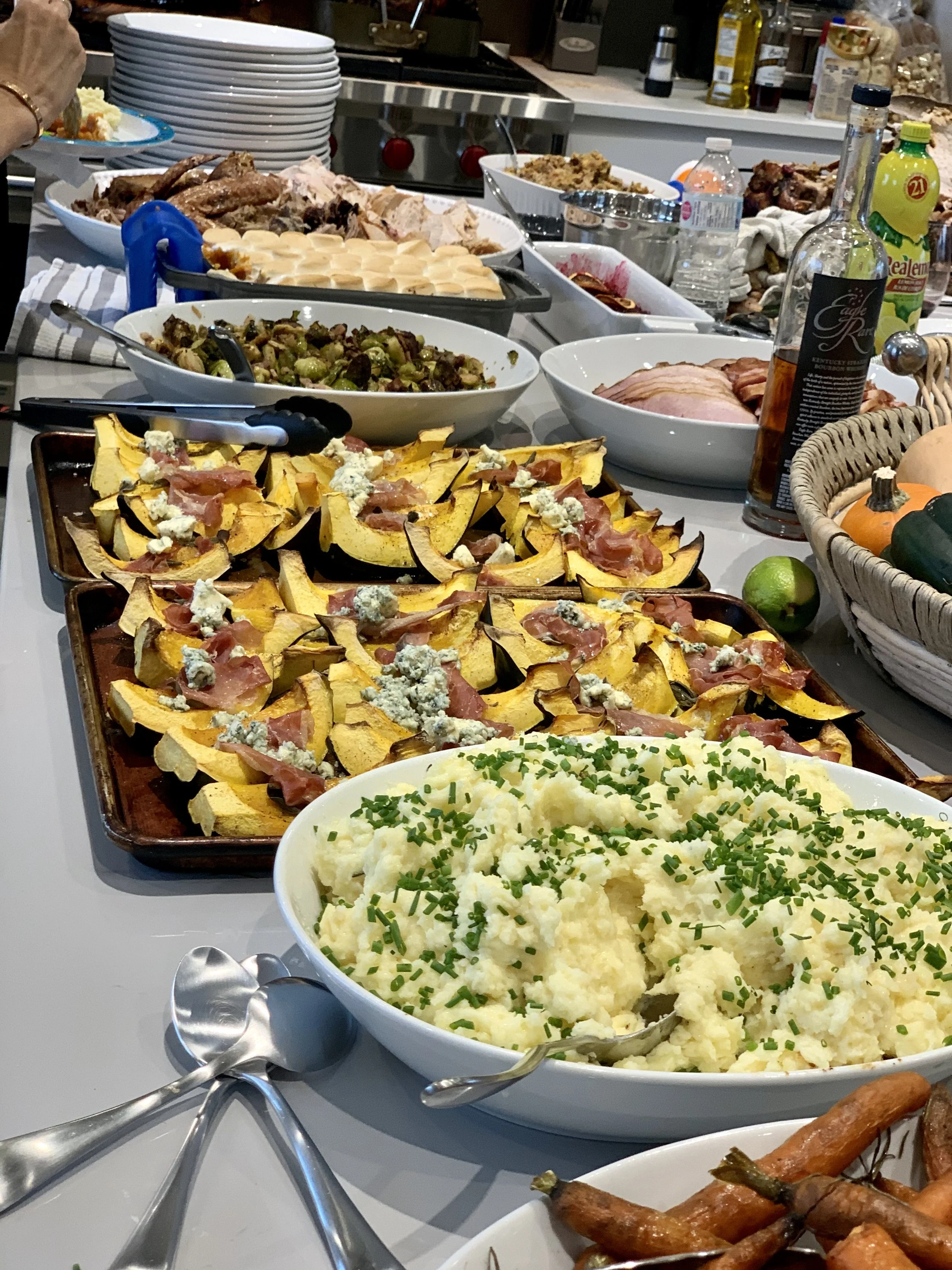 A variety of dishes on a table, including mashed potatoes topped with green herbs, roasted vegetables, sliced meats, stuffed squash or zucchini, and assorted sides, with additional food and drinks in the background.