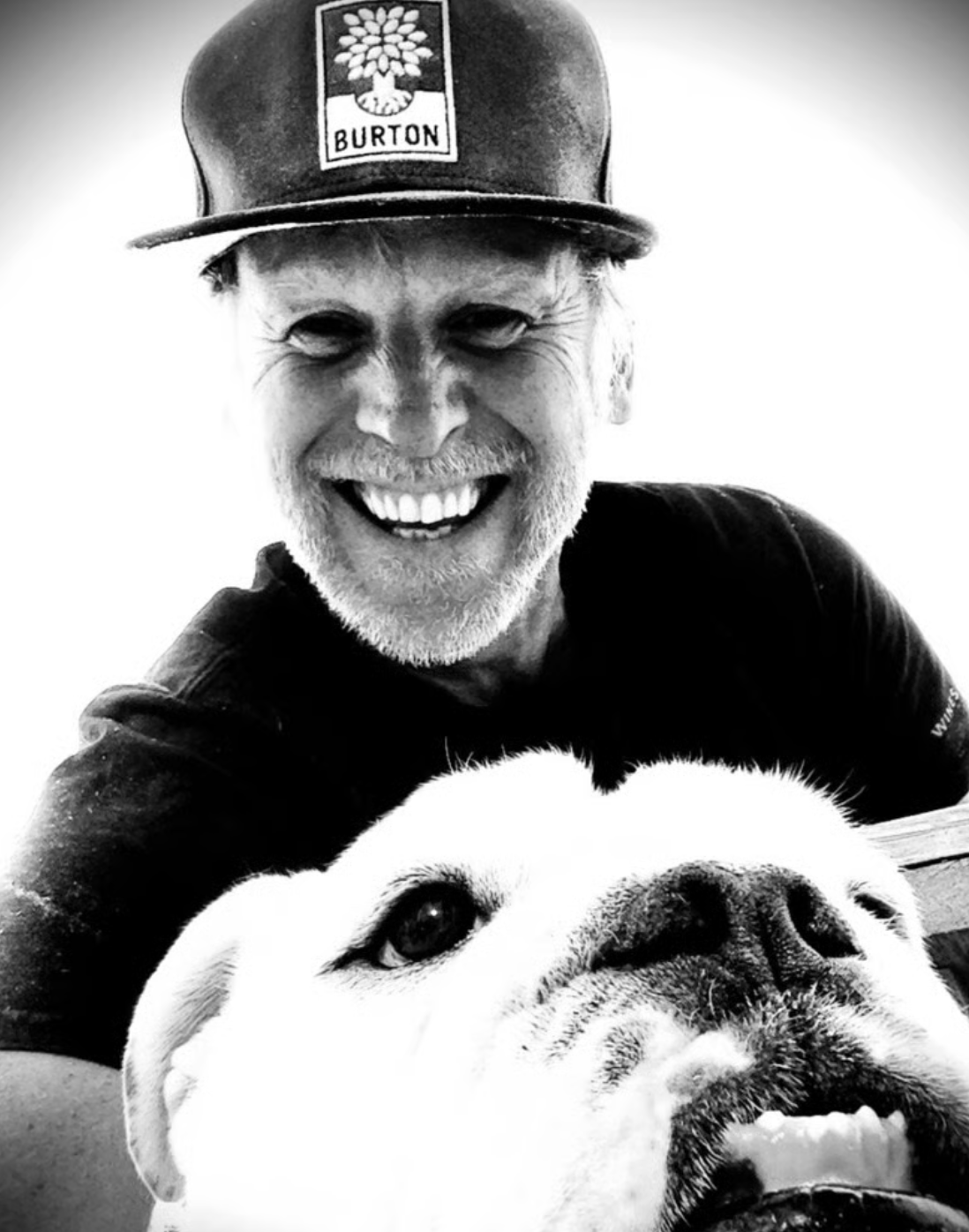 A smiling man with a beard wearing a baseball cap and a dark shirt, lying on his stomach with a Bulldog dog in front of him, in black and white.