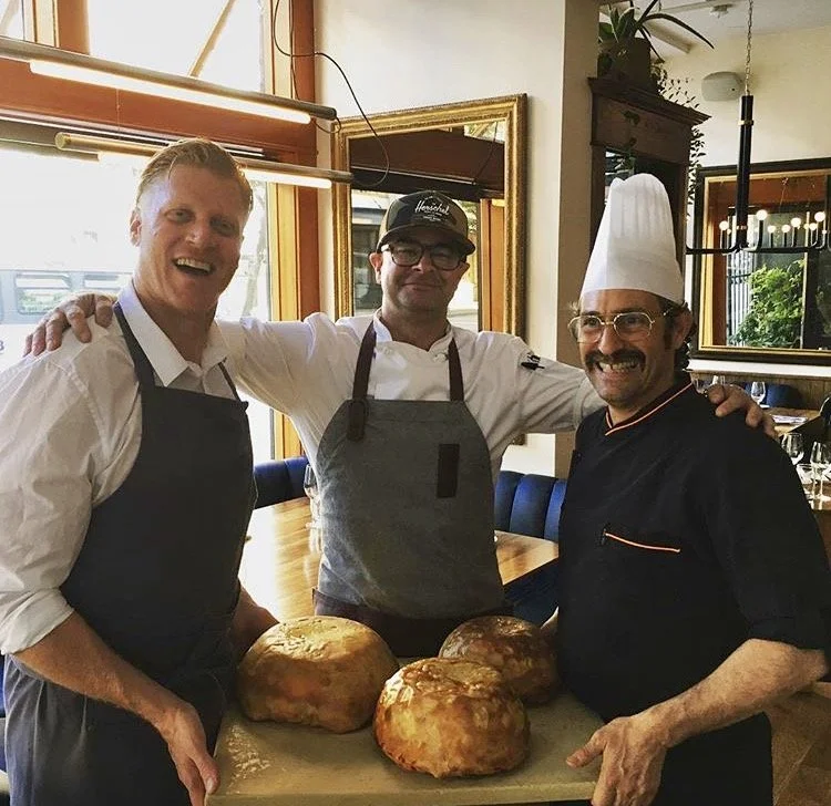 Three male chefs smiling, posing behind three loaves of bread on a table in a restaurant setting.