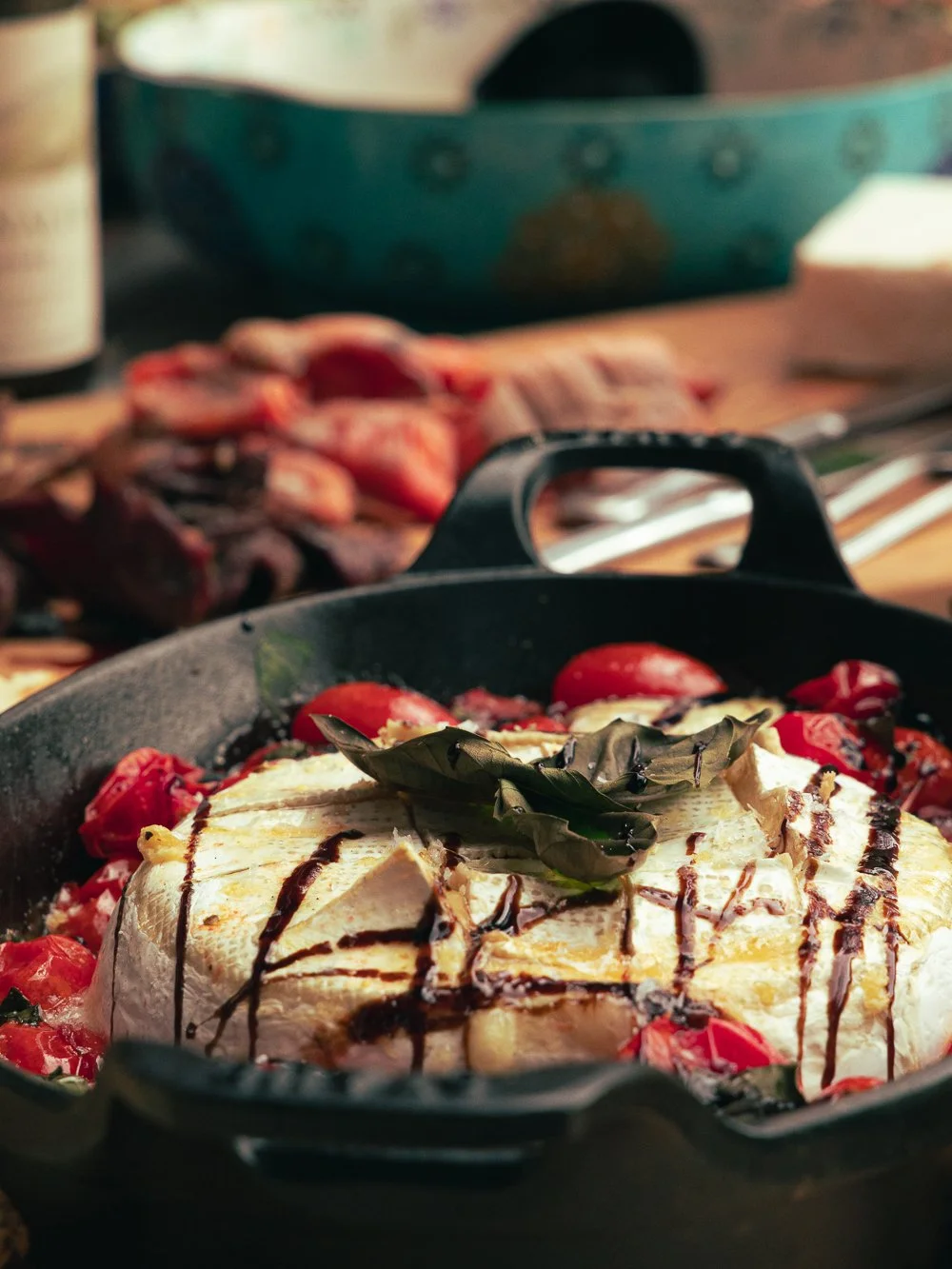 Baked pasta dish with cheese, cherry tomatoes, basil, and balsamic glaze in a black skillet on a wooden table, with a blurred background of other ingredients and utensils.