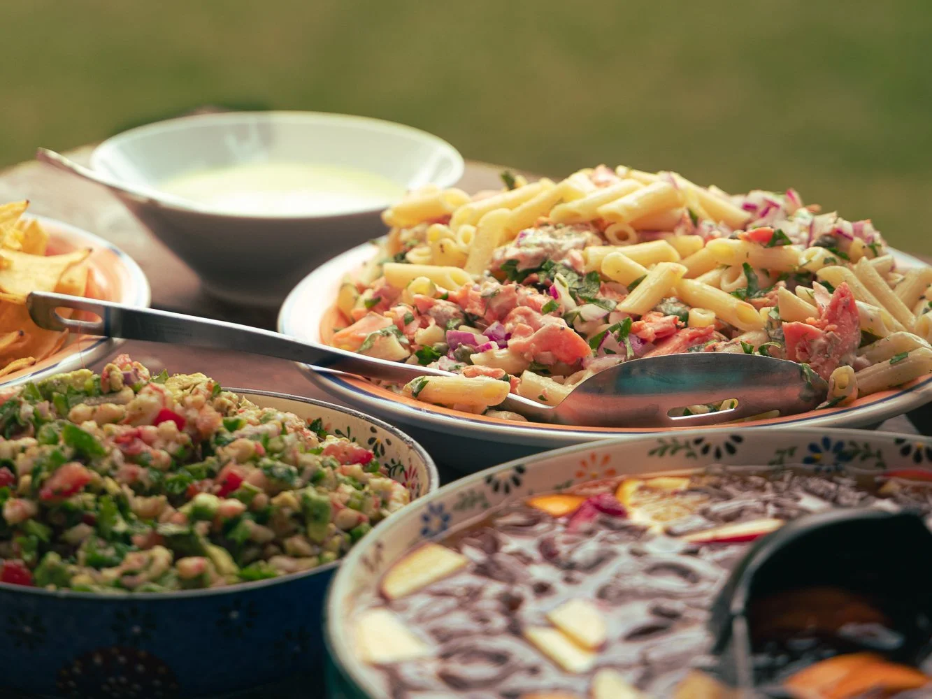 Assorted bowls of pasta salad, vegetable salad, and dipping sauces on a picnic table outdoors.