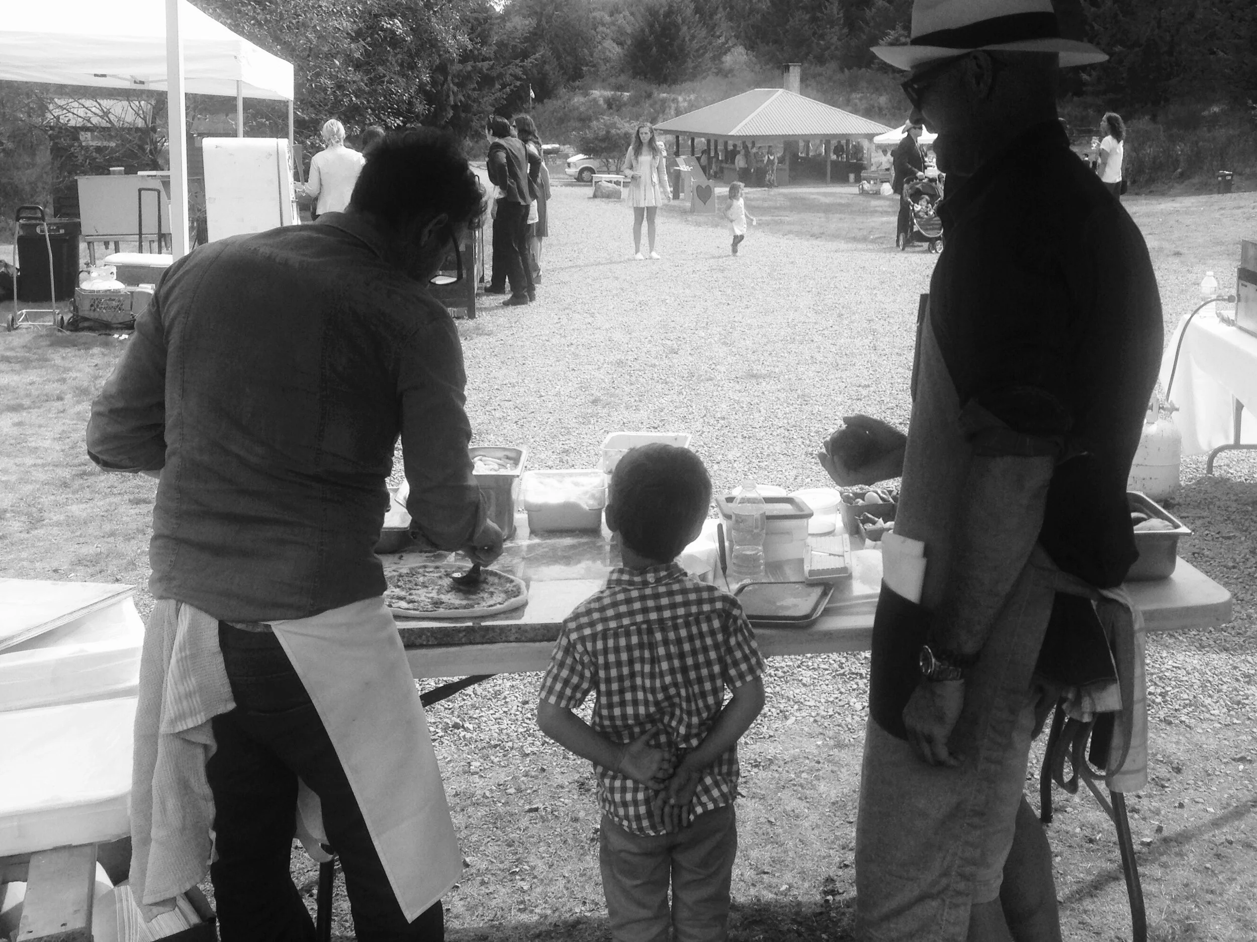 People gathered at an outdoor event, including a young boy in a checkered shirt, two adults wearing aprons, and another two people under an umbrella. In the background, there are more individuals, tents, and a pavilion in a park setting.