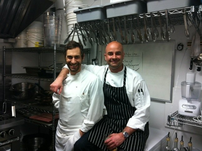 Two male chefs in white uniforms and striped apron sitting in a professional kitchen, smiling at the camera.